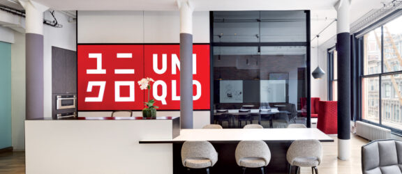 Modern commercial interior featuring a minimalist reception area with a sleek white desk, gray upholstered seating, and bold red graphic art. Overhead, geometric black lighting fixtures contrast with the exposed white ceiling, accentuating the space's contemporary aesthetic.