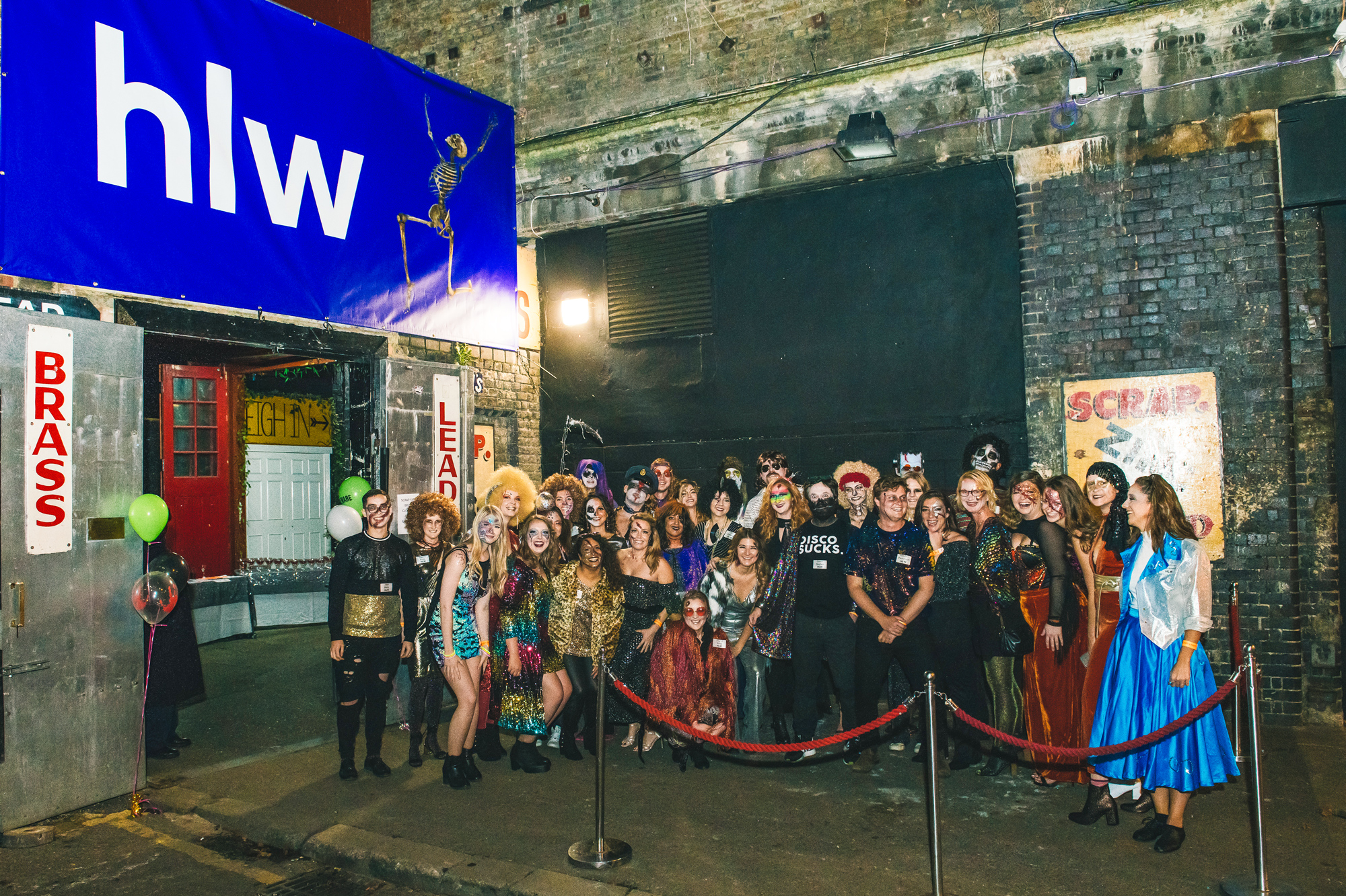 Large group of smiling people in diverse and colorful costumes gathered for a festive event, with a 'hlw' sign and urban decor setting the backdrop.