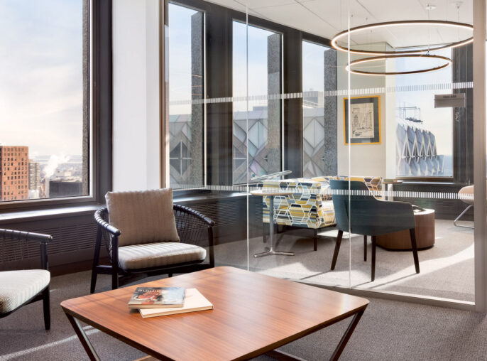 Modern office space with expansive windows offering city views, complemented by geometric furnishings and circular ceiling light fixtures. The neutral palette is accented by a pop of color from wall art and textiles.