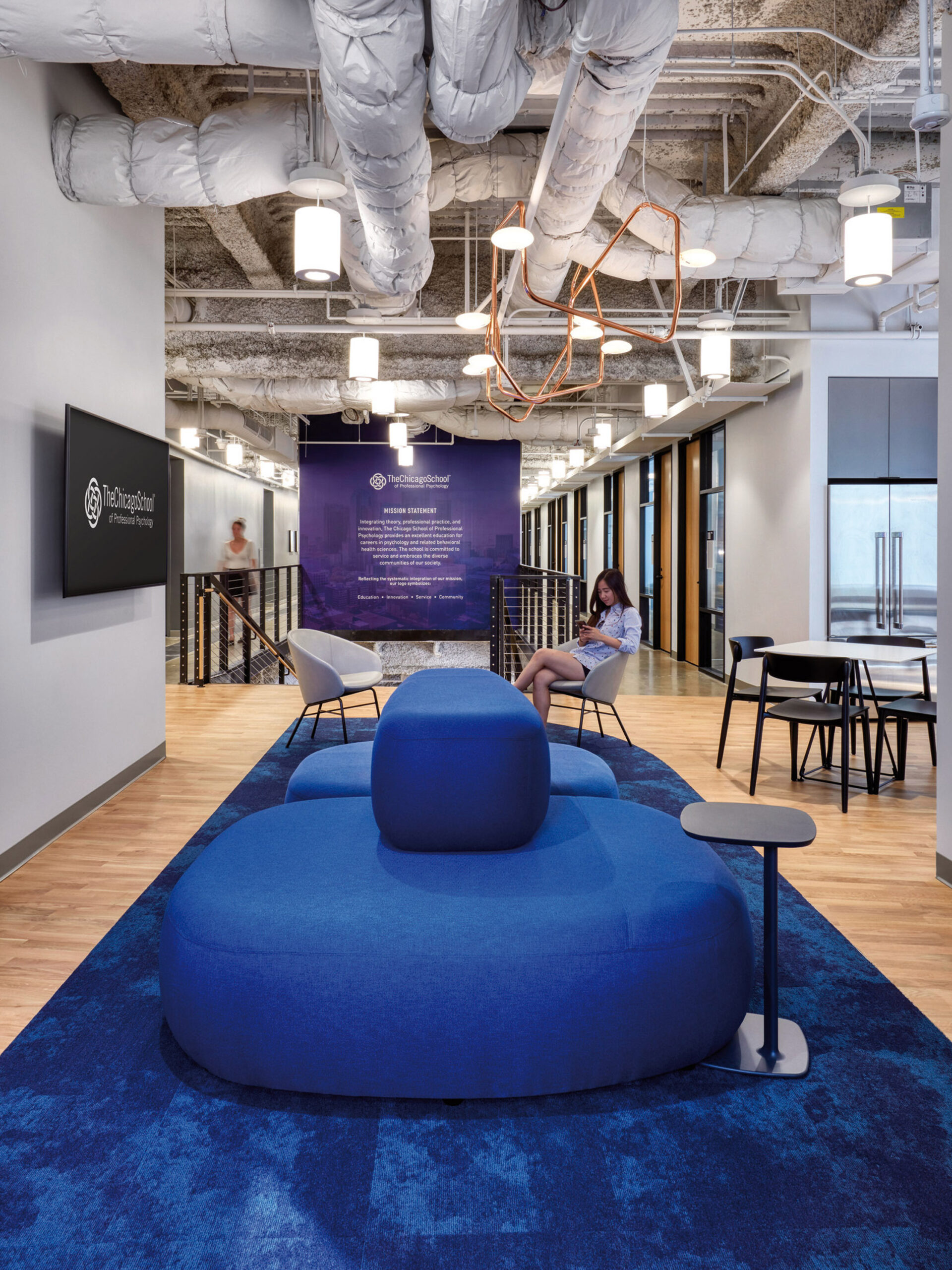 Open-plan office space featuring exposed ductwork and pendant lighting, with a vibrant blue area rug anchoring modular seating. Neutral walls showcase company branding, creating a contemporary, collaborative atmosphere.