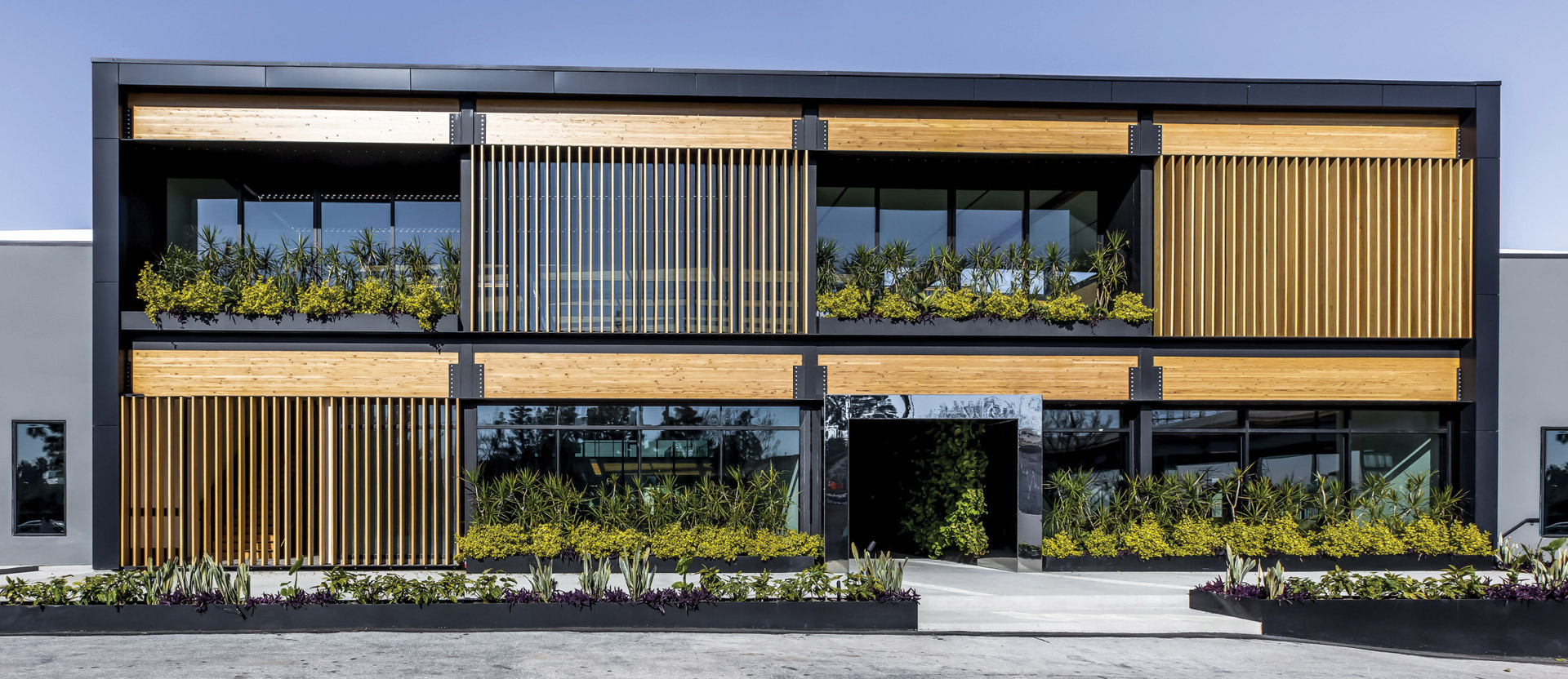 Modern two-story commercial building featuring a balanced mix of materials with horizontal wood paneling, vertical metal louvers, and generous windows framed by black trim. Lush green planters add a natural touch to the sleek, contemporary design.