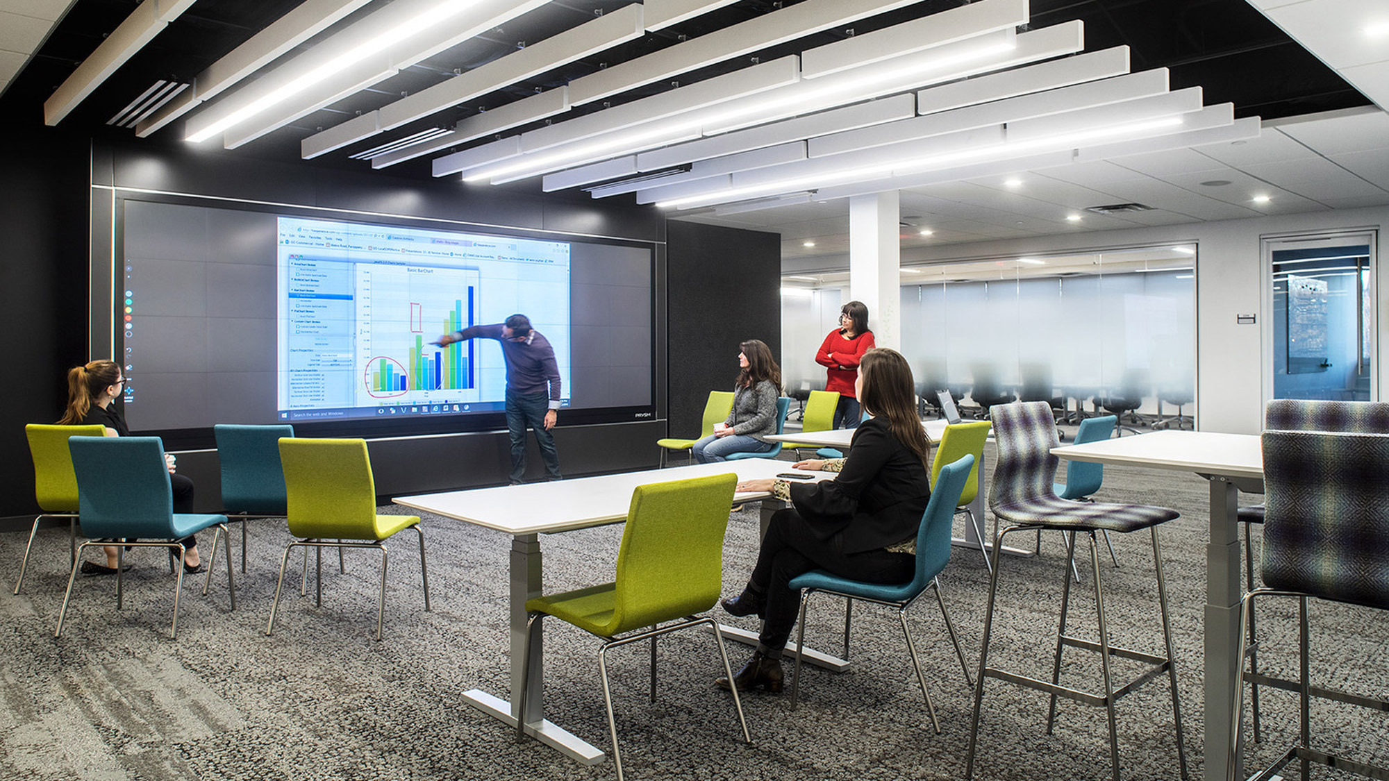 Modern office meeting area with a large digital presentation screen. Colorful chairs in green and blue hues contrast with the monochromatic carpet. Overhead, linear lights complement the sleek, contemporary vibe of the workspace.