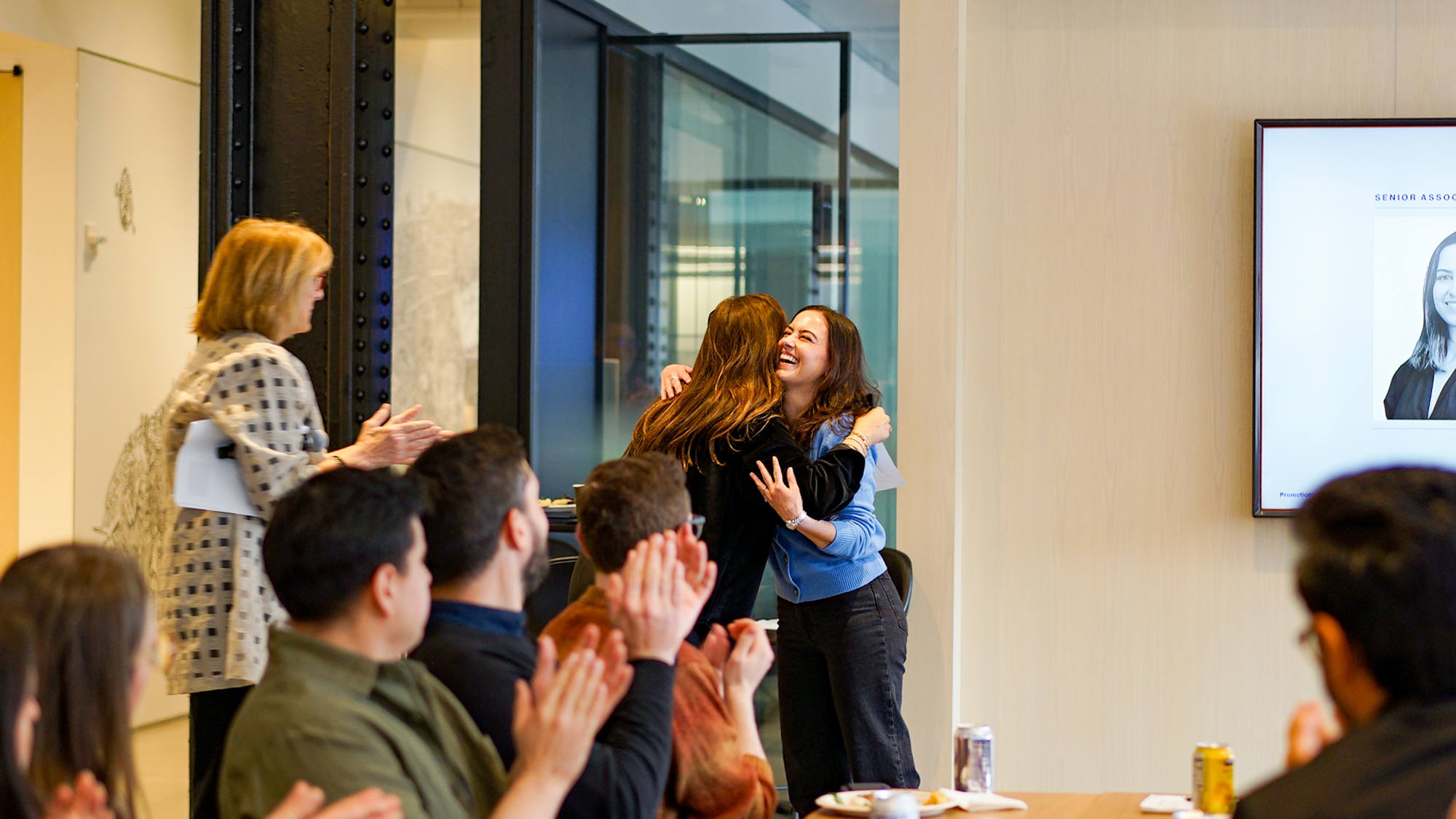 A joyous moment in a corporate office setting as two colleagues embrace in a congratulatory hug with a backdrop of applauding team members and a presentation screen, capturing the essence of professional recognition and team spirit.