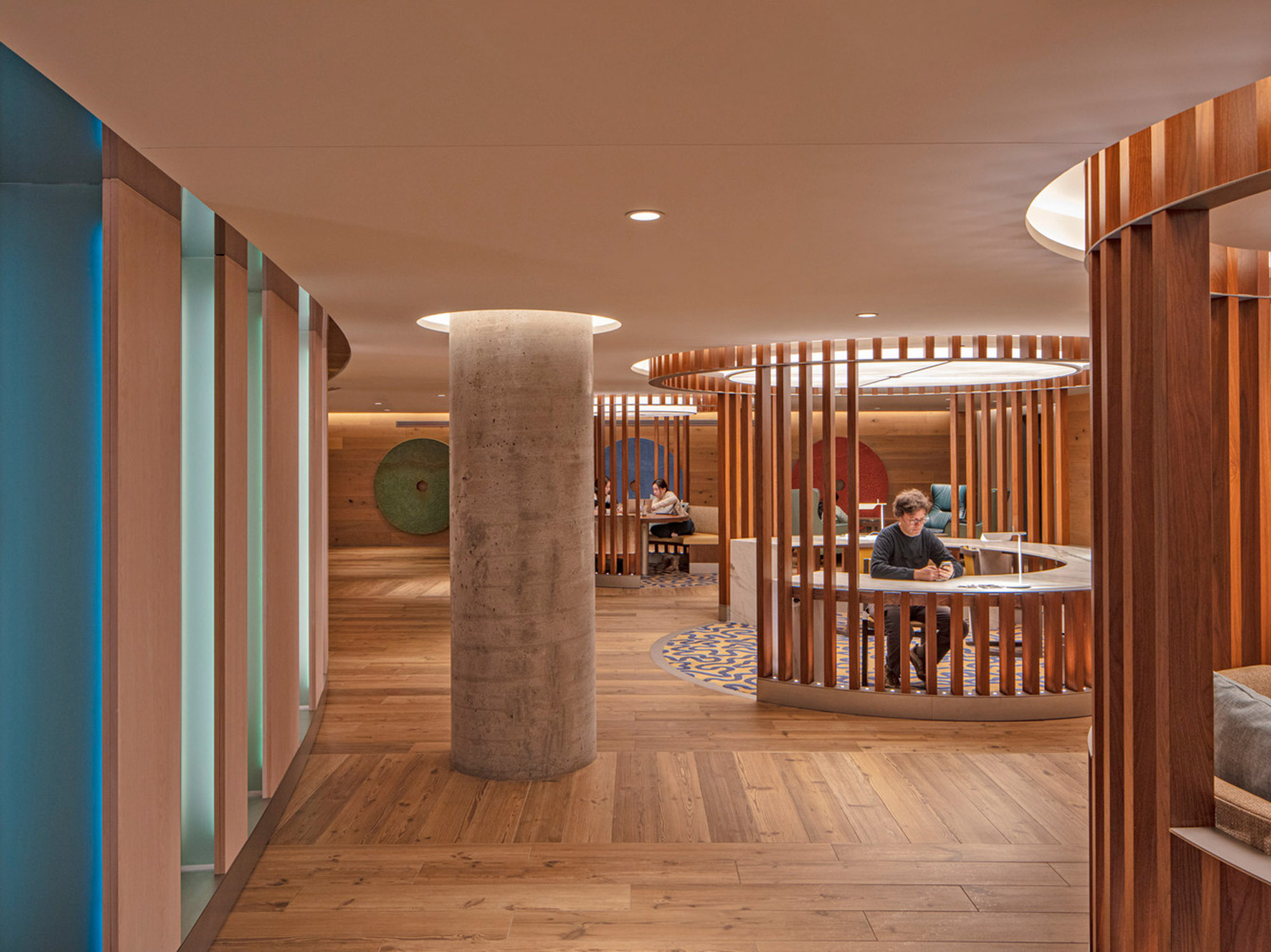 Modern office interior with warm wooden slats creating semi-private workspaces, complemented by subdued lighting and a central exposed concrete column. Geometric carpet patterns and wall art add vibrancy to the space.