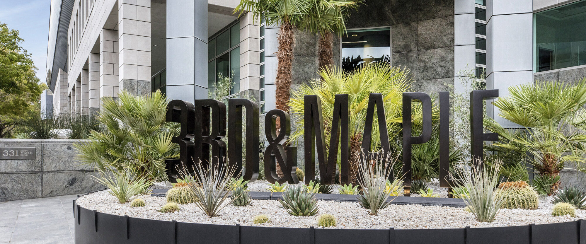 Modern commercial landscape featuring bold 3D letter signage amidst an array of desert landscaping elements, such as agave and yucca plants, set against a building with reflective glass and granite finishes.