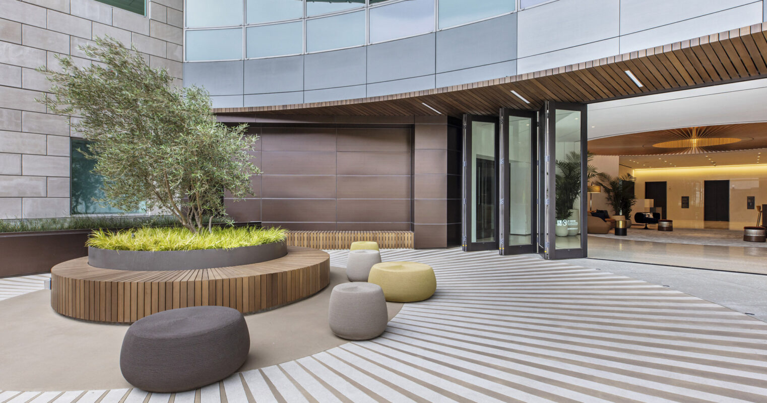 Modern office lobby with a minimalist design featuring a combination of natural and geometric elements. A central, circular wooden seating area incorporates greenery, surrounded by parallel stone tiles and contrasting soft, rounded poufs. Clean lines and a glass facade allow natural light to accentuate the space's textures and materials.