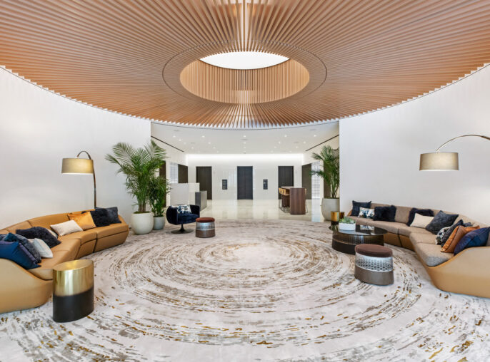 Modern lobby with a concentric patterned marble floor leading to tan sofas accented with blue and gray pillows. Curved wooden slat ceiling with central lighting feature, flanked by tall potted plants and contemporary floor lamps, conveys a warm, inviting atmosphere.