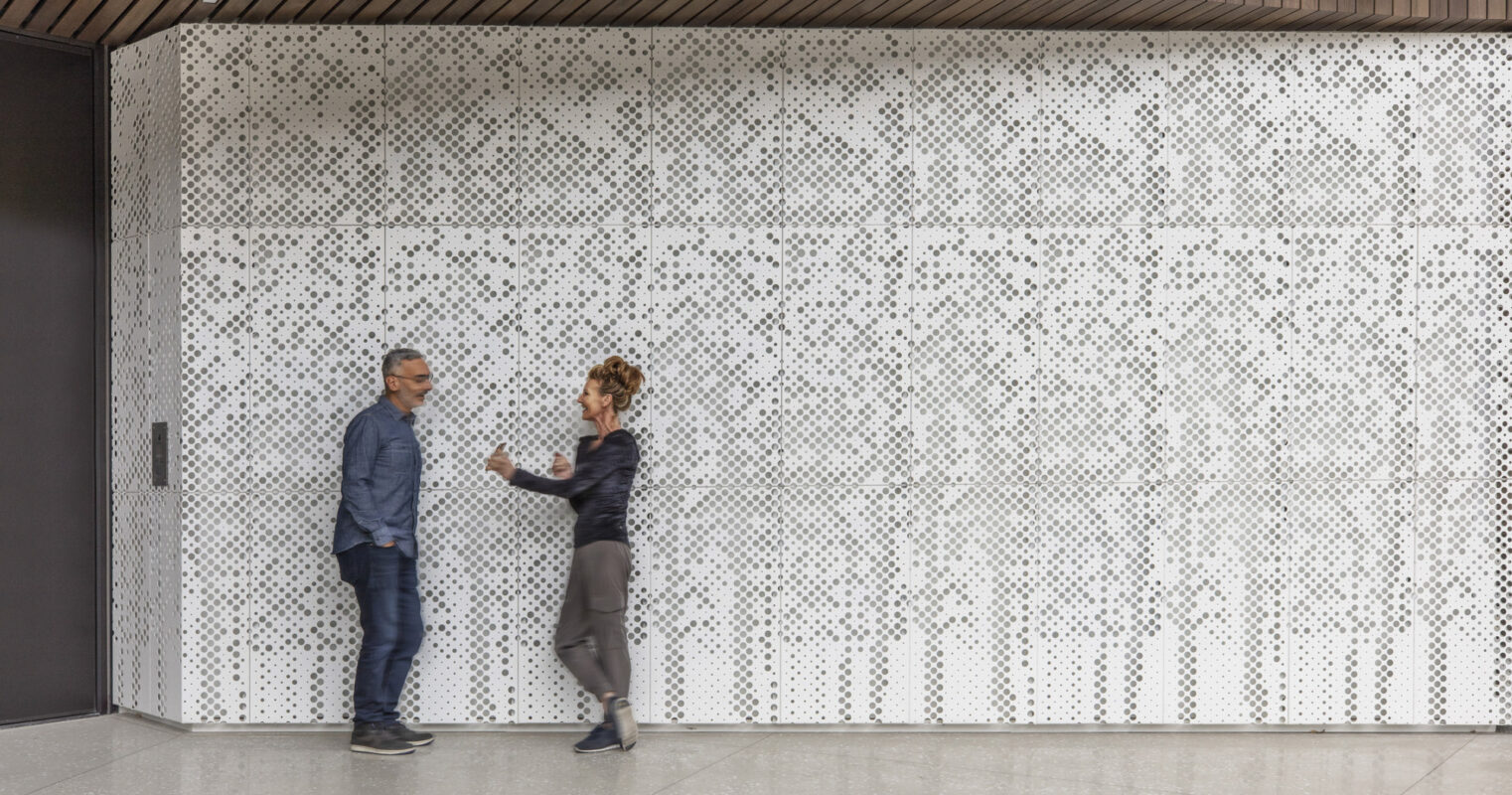 Two individuals converse in a modern hallway featuring a geometric-patterned perforated metal screen wall, providing both privacy and light diffusion, accented by a warm wood ceiling and minimalist design.