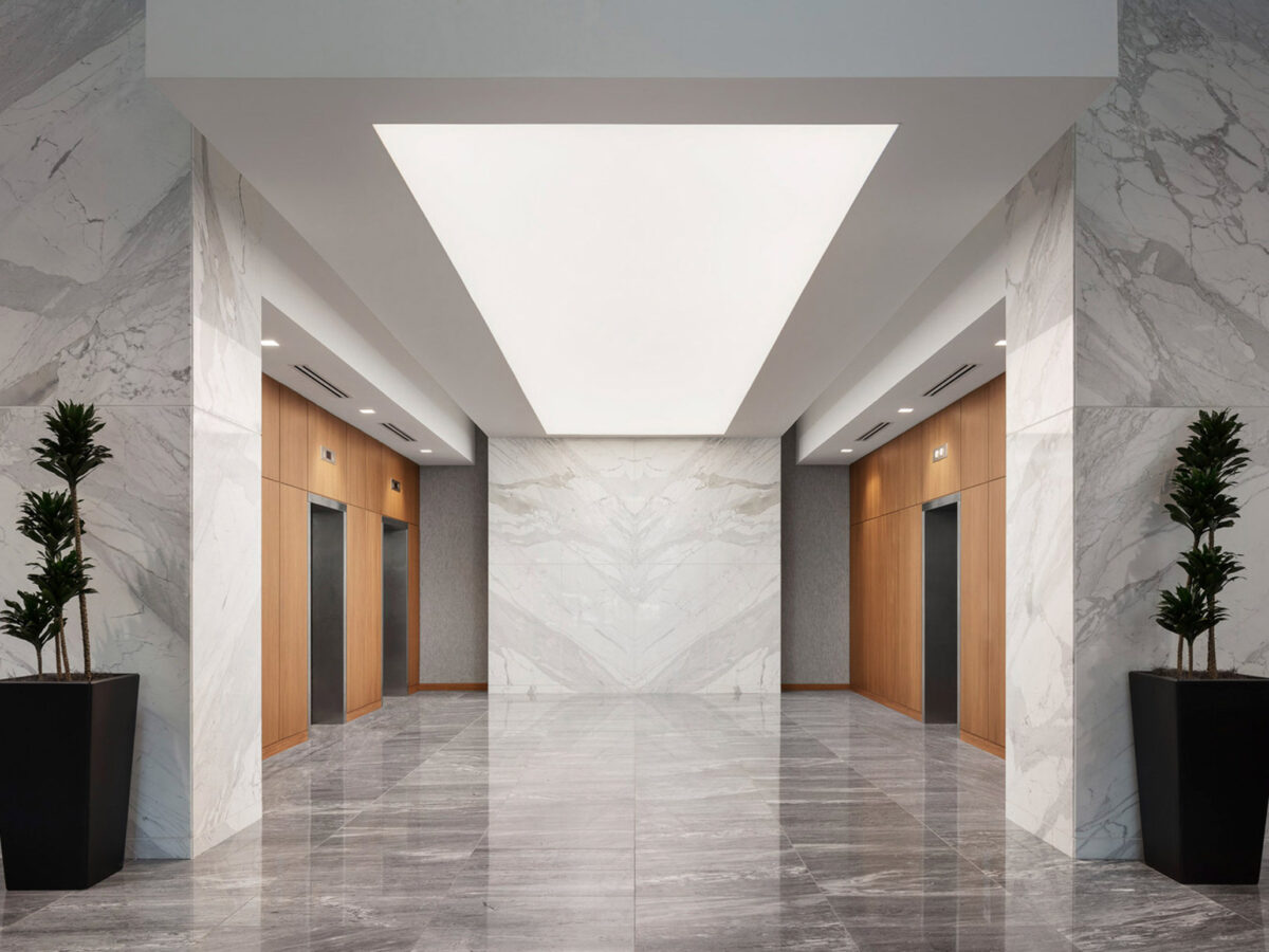 Modern lobby with a geometric ceiling light fixture casting a soft glow on the gleaming marble floor. The walls, adorned in matching marble, lead to wooden doors, flanked by potted plants, creating a harmonious blend of natural and contemporary design elements.