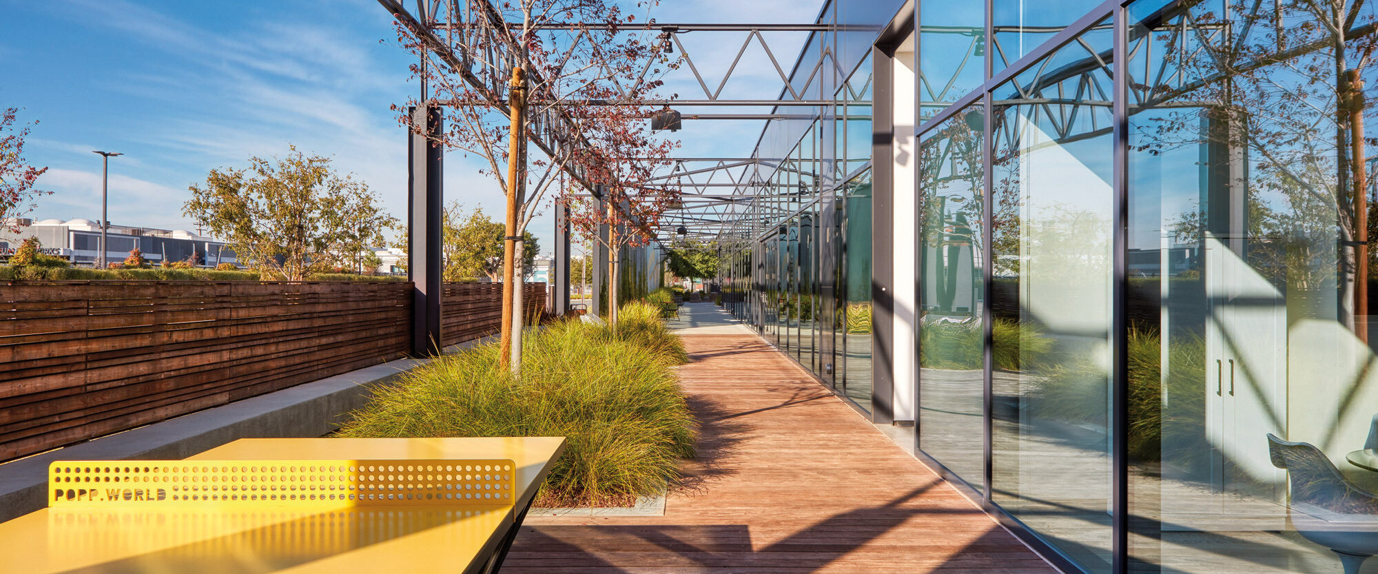 Lush outdoor corridor with a wooden pathway flanked by full-glass walls and green landscaping. Structural columns support a modern pergola, casting geometric shadows. A vibrant yellow bench adds a pop of color to the serene, natural setting.