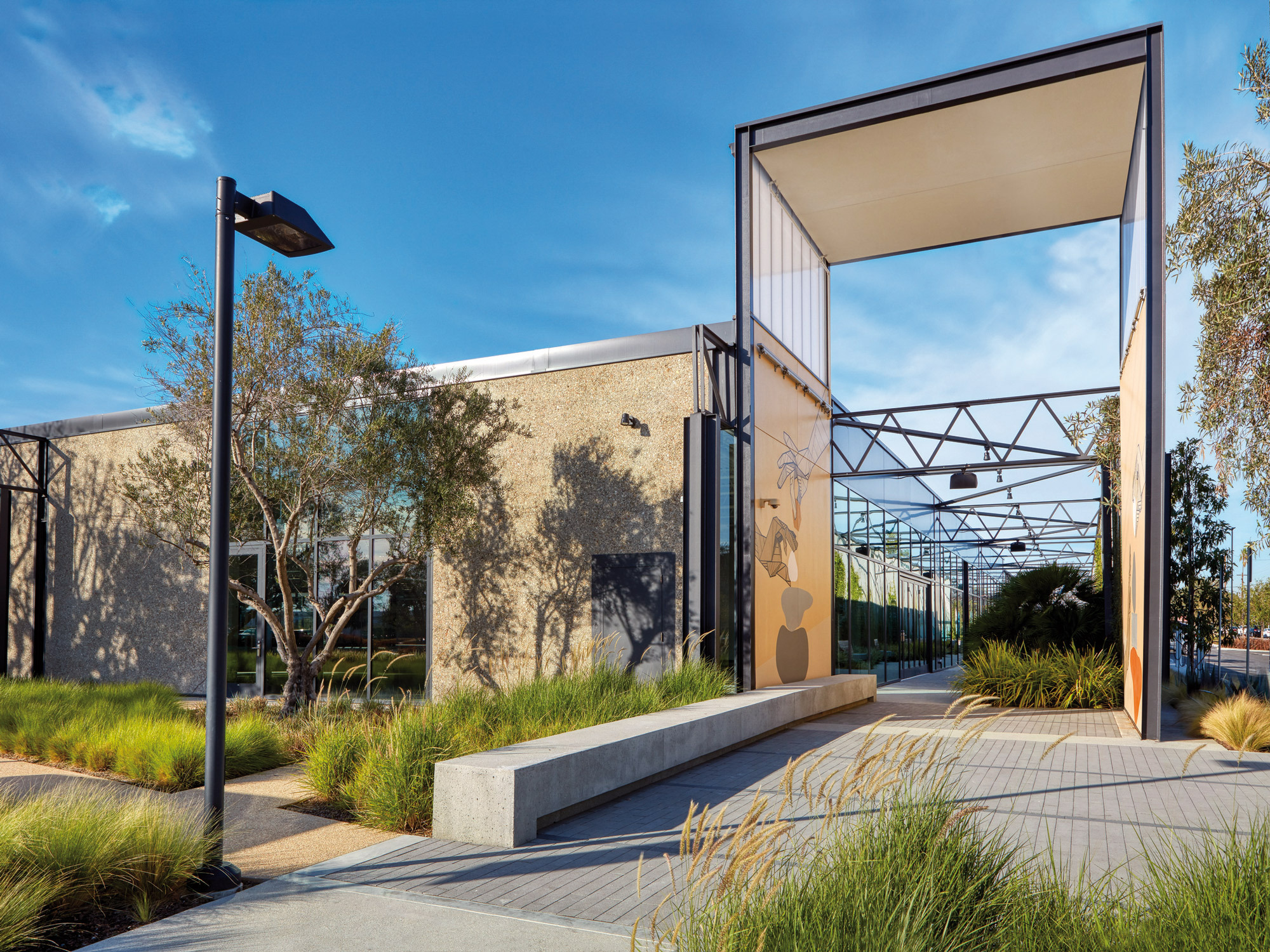 Modern commercial building entrance with full-height glass doors, framed by sleek steel beams and a cantilevered canopy. Landscaped with ornamental grasses and concrete steps, it blends functional design with natural elements.