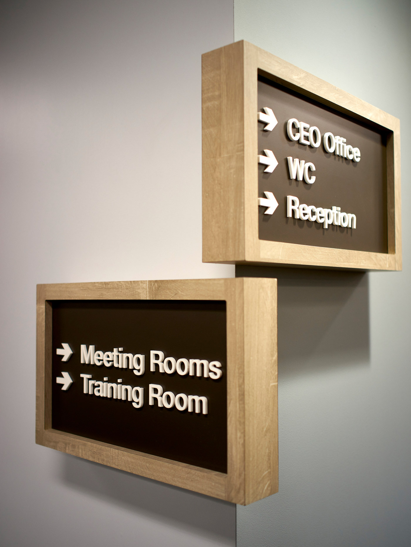 Wall-mounted wooden signage featuring backlit frosted glass panels displays directional information for a CEO office, WC, Reception, Meeting Rooms, and Training Room, with white arrow symbols and text.