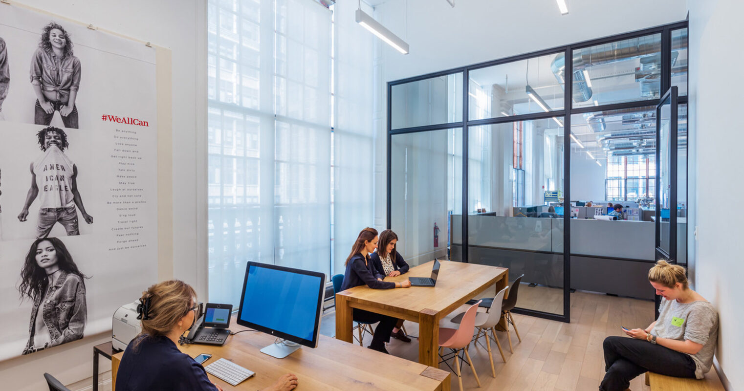 Modern office space featuring a minimalist wooden table with workers on laptops, surrounded by white walls adorned with large format art, expansive windows providing ample natural light, and clear glass partitionings enhancing the open-plan design aesthetic.
