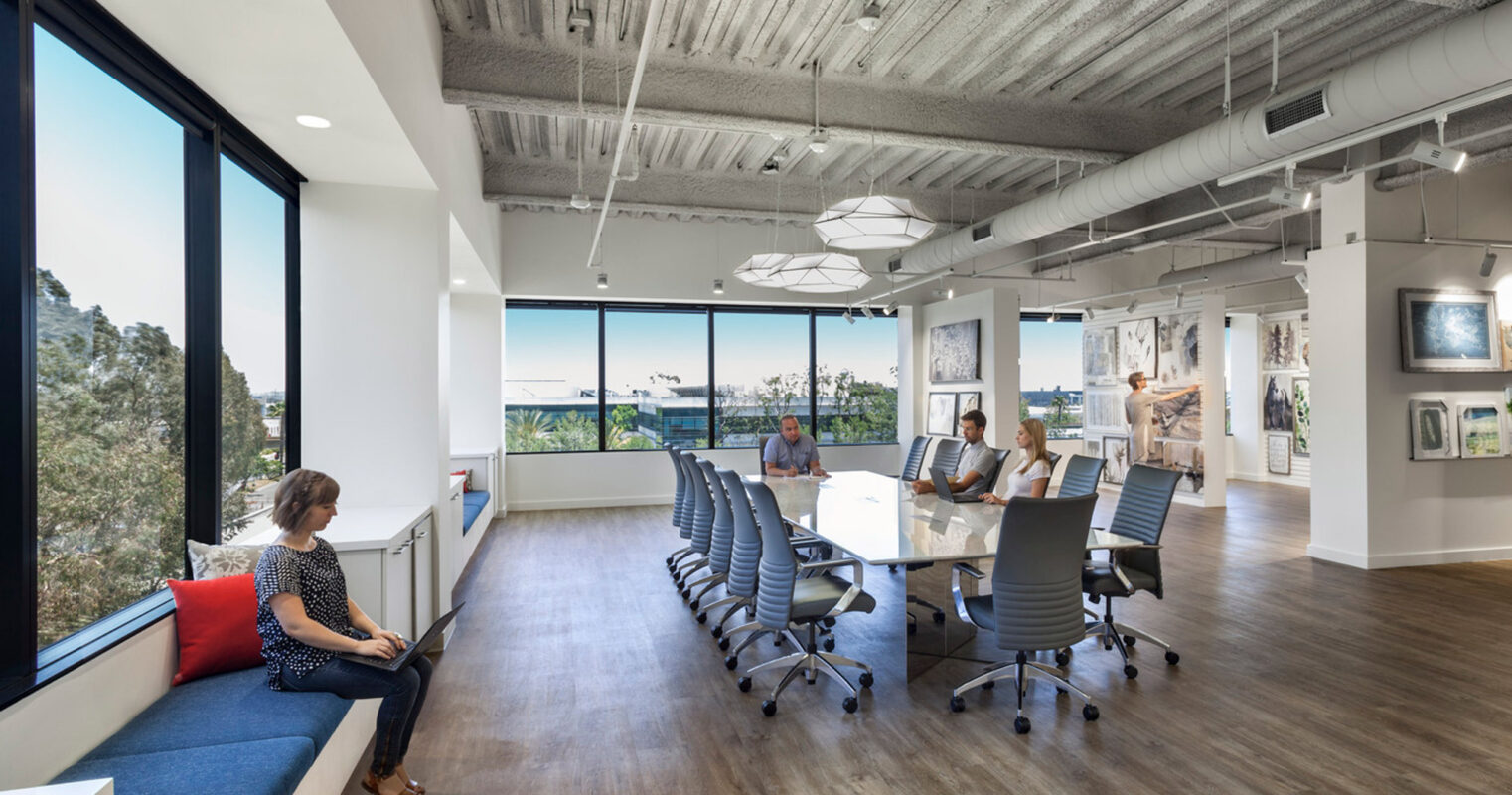 Modern office space boasting exposed concrete ceiling and ductwork, with floor-to-ceiling windows offering natural light and a view of greenery. Sleek, linear lighting fixtures complement a long conference table, while colorful artwork adds visual interest along a neutral-toned wall.