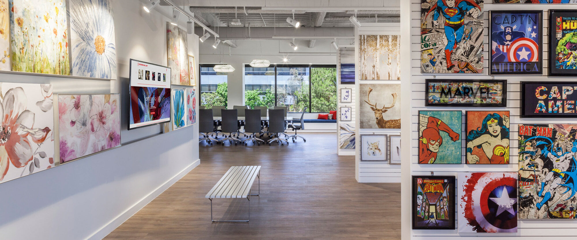 Bright and spacious gallery featuring modern artwork. Exposed ductwork contrasts with white walls adorned with vibrant paintings and comic-themed pieces. Sleek bench and parquet floor complement the room's contemporary aesthetic.