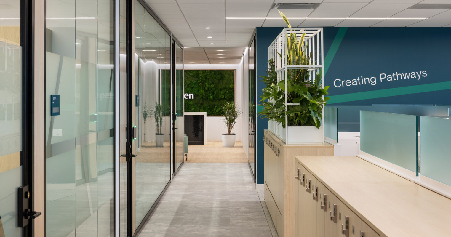 Modern office corridor with sleek glass partitions, reflecting a functional yet aesthetically pleasing design. A neutral color palette is complemented by a vibrant teal accent wall, inscribed with motivational text. Potted greenery adds a touch of nature, enhancing the workspace's ambiance.