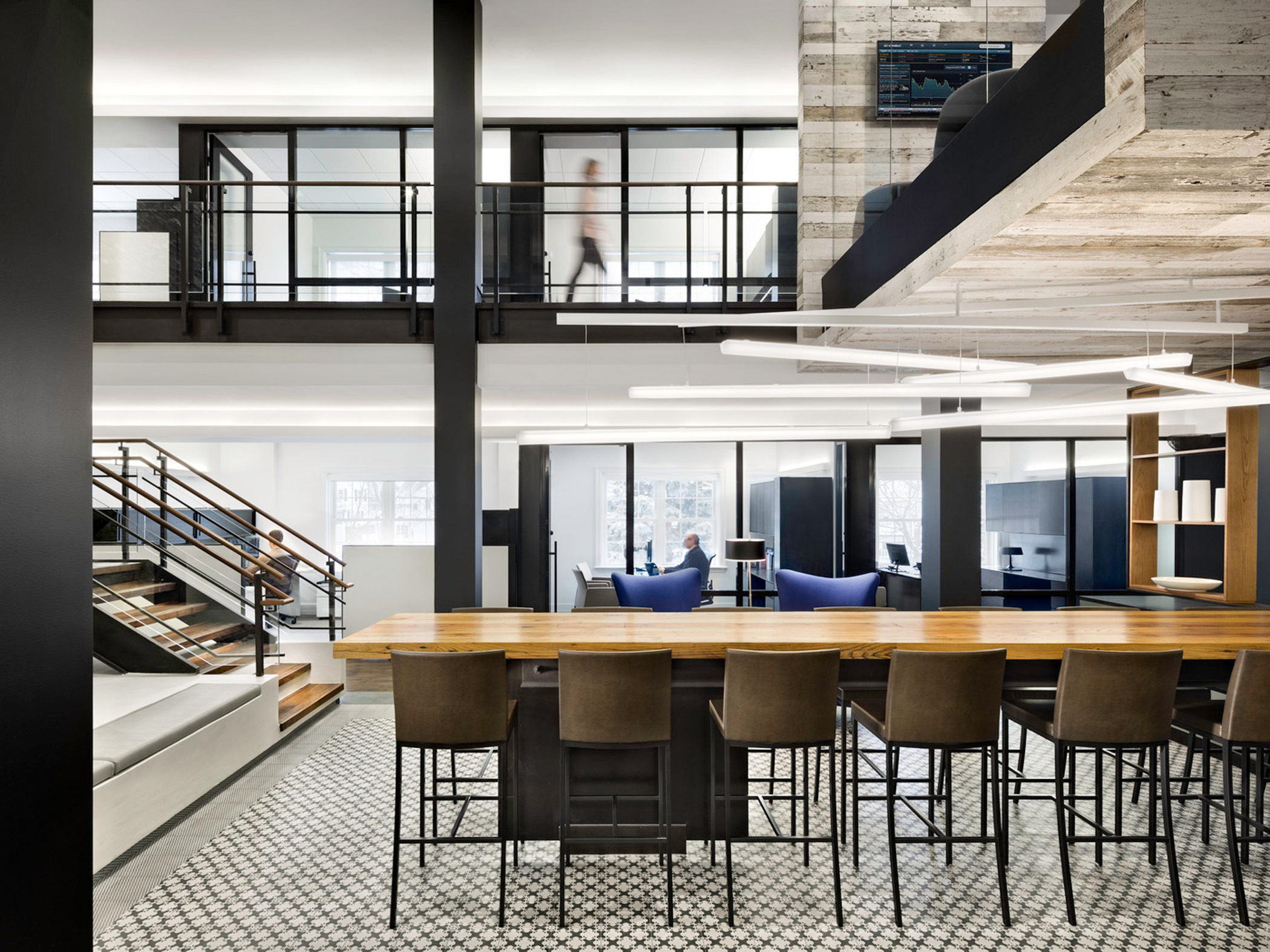 Modern office interior showcasing an open workspace with wood-topped bar-style seating, black steel-frame mezzanine, geometric patterned flooring, and suspended linear lighting fixtures. A person walks across the transparent walkway, adding human scale to the minimalist design.