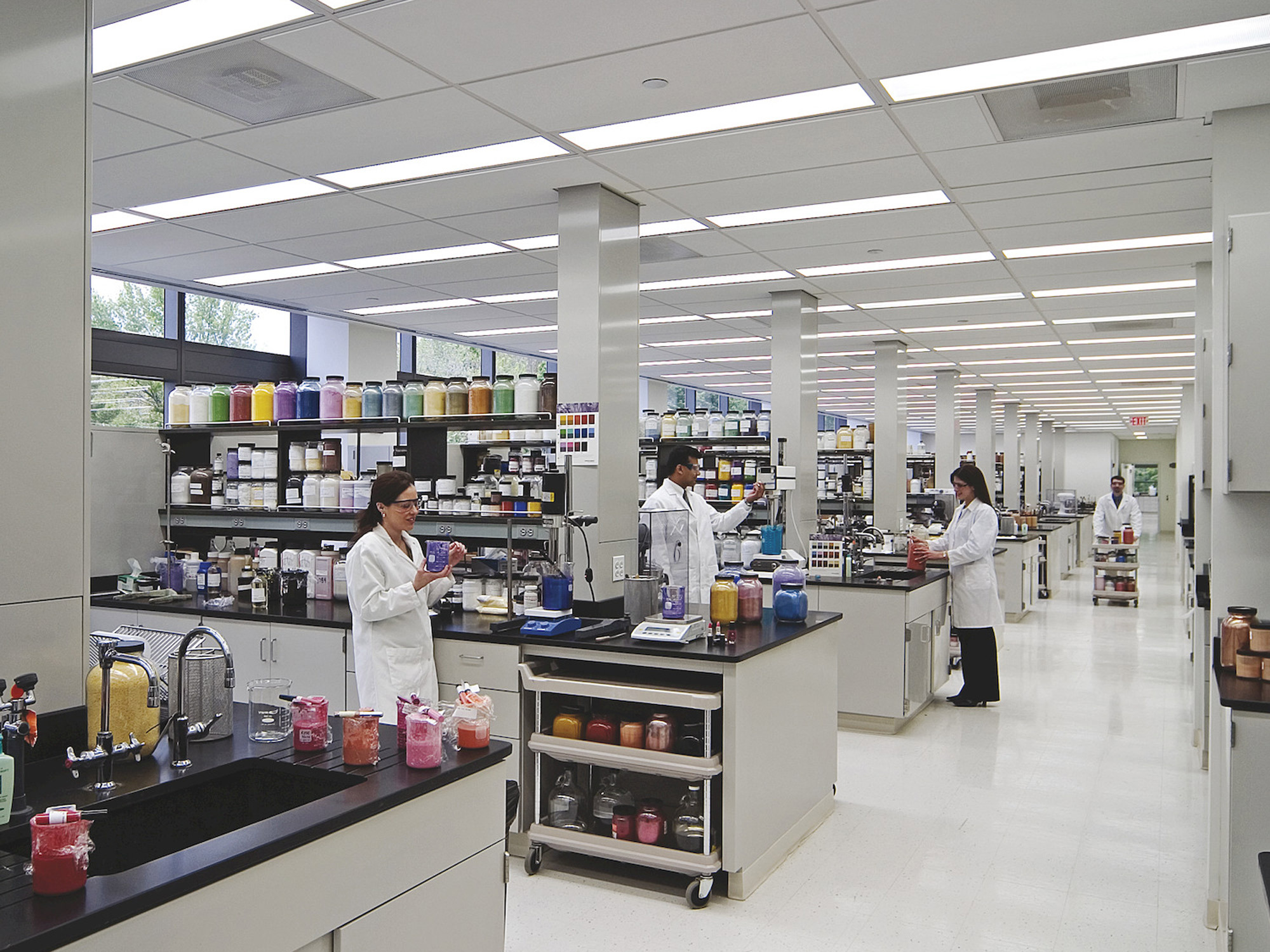 Spacious laboratory interior with streamlined white countertops; equipped with scientific apparatus, glassware, and labeled material containers. Scientists in lab coats engaged in research activities, enhancing the space's functional design highlighting cleanliness and organization for efficient workflow.