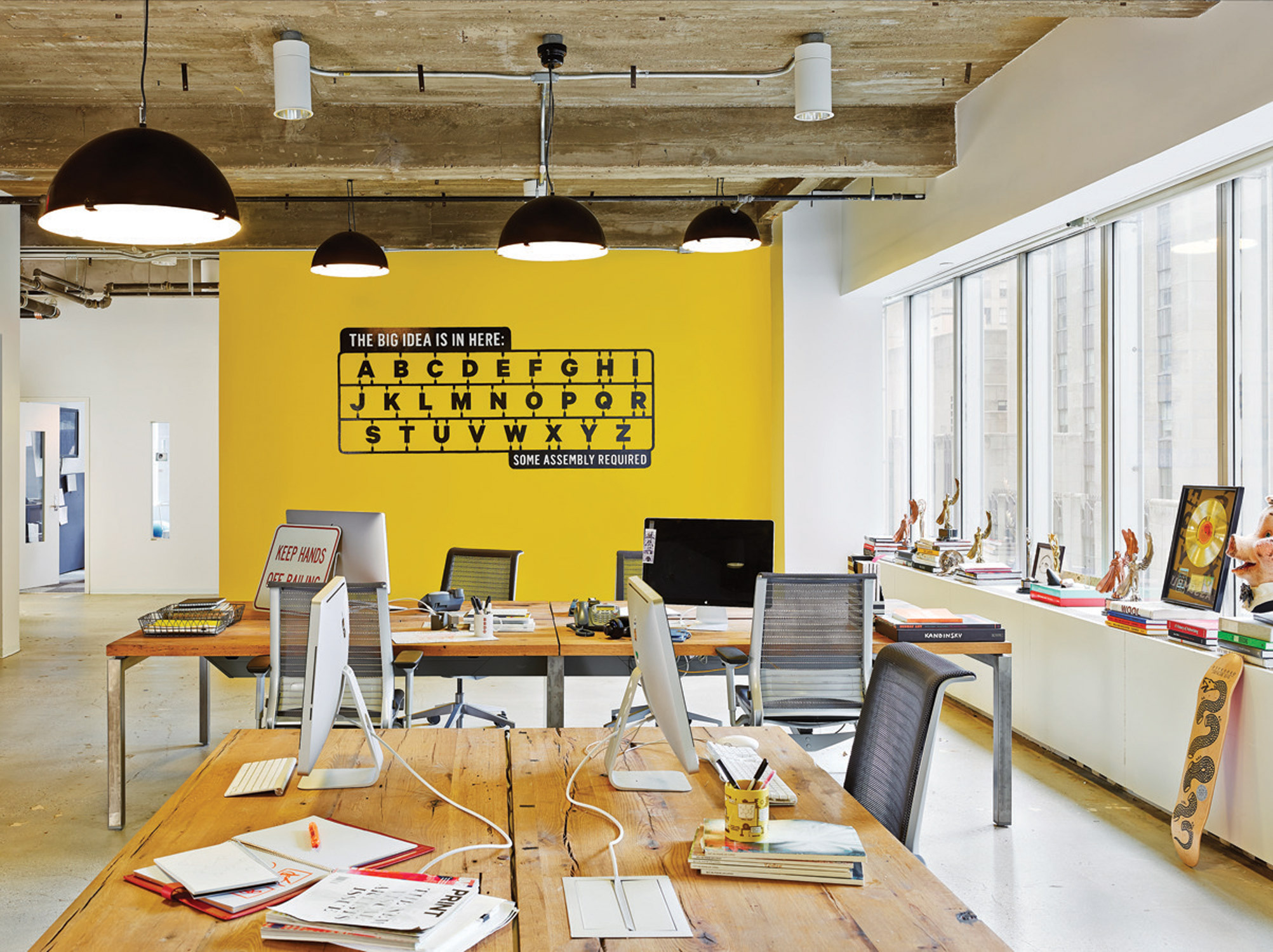 Lively office space with a yellow accent wall featuring typographic art, surrounded by workstations with exposed concrete ceilings.