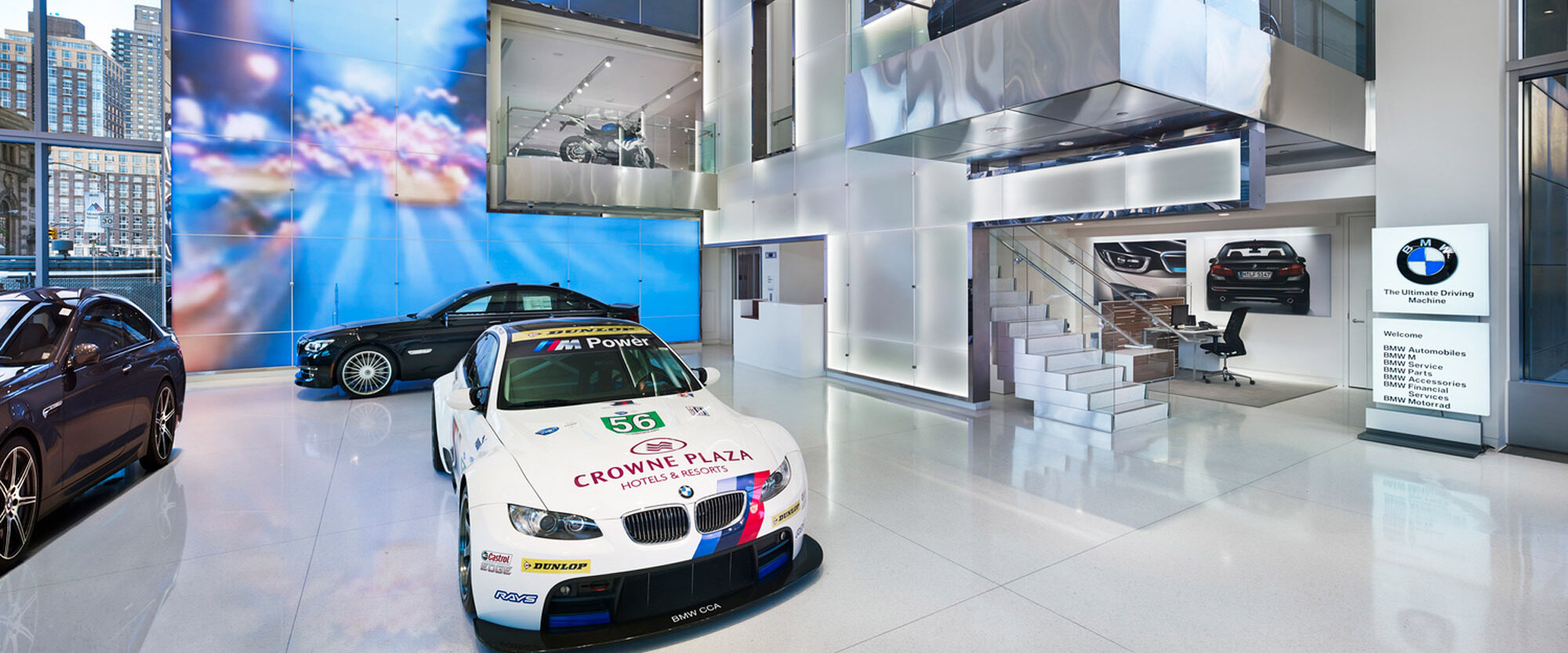 Spacious automotive showroom with gleaming white floors, full-height windows, and multiple cars on display, including a prominent race car. Two-story glass partitions enhance the modern, airy ambiance, while sleek metallic staircases offer views to the second level.