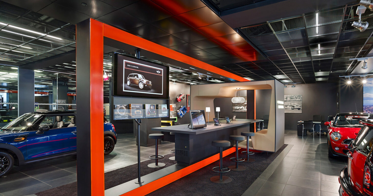Modern car showroom with vibrant blue and orange accents, featuring sleek desks, interactive display screens, and a selection of luxury vehicles on a polished concrete floor, all under strategic lighting that highlights the cars’ designs.