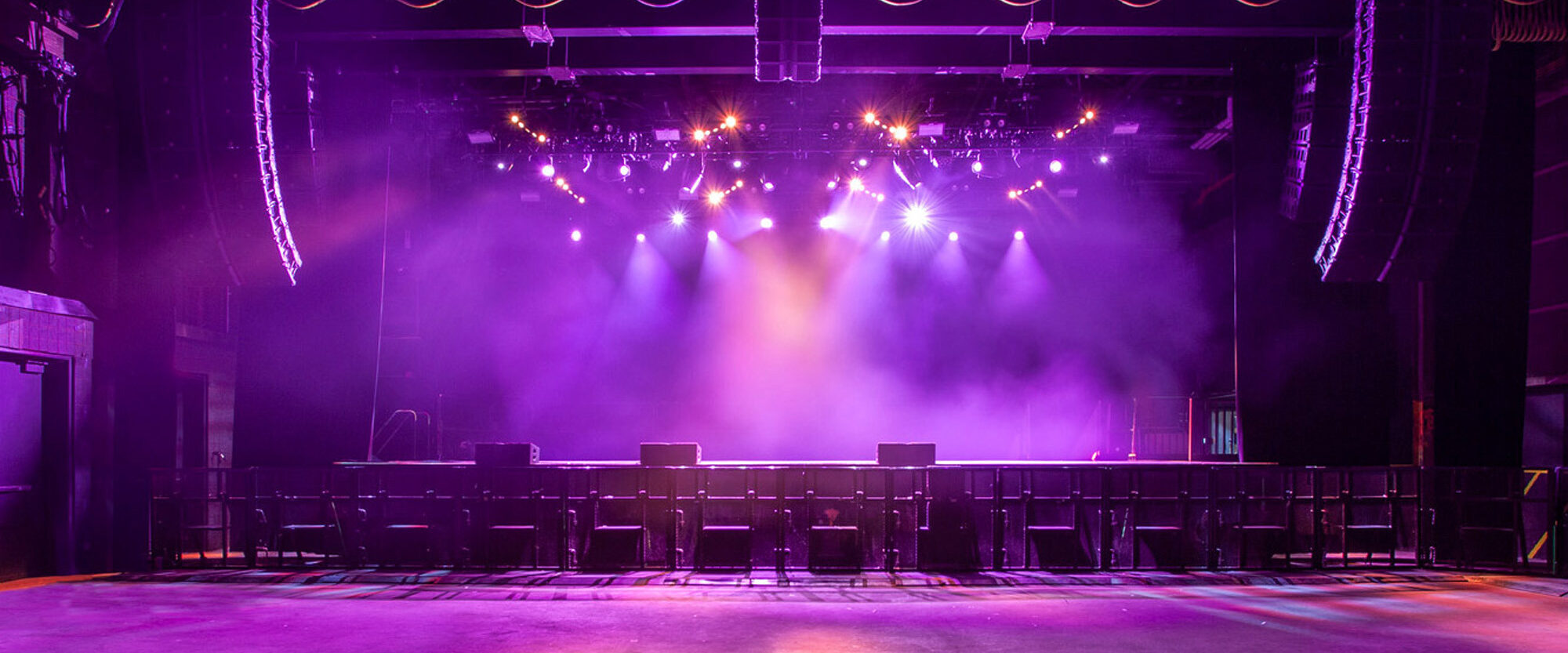 Vibrant stage lighting illuminates an empty concert venue with rows of chairs facing a stage, casting dramatic shadows and showcasing a contemporary design melding performance with aesthetics.