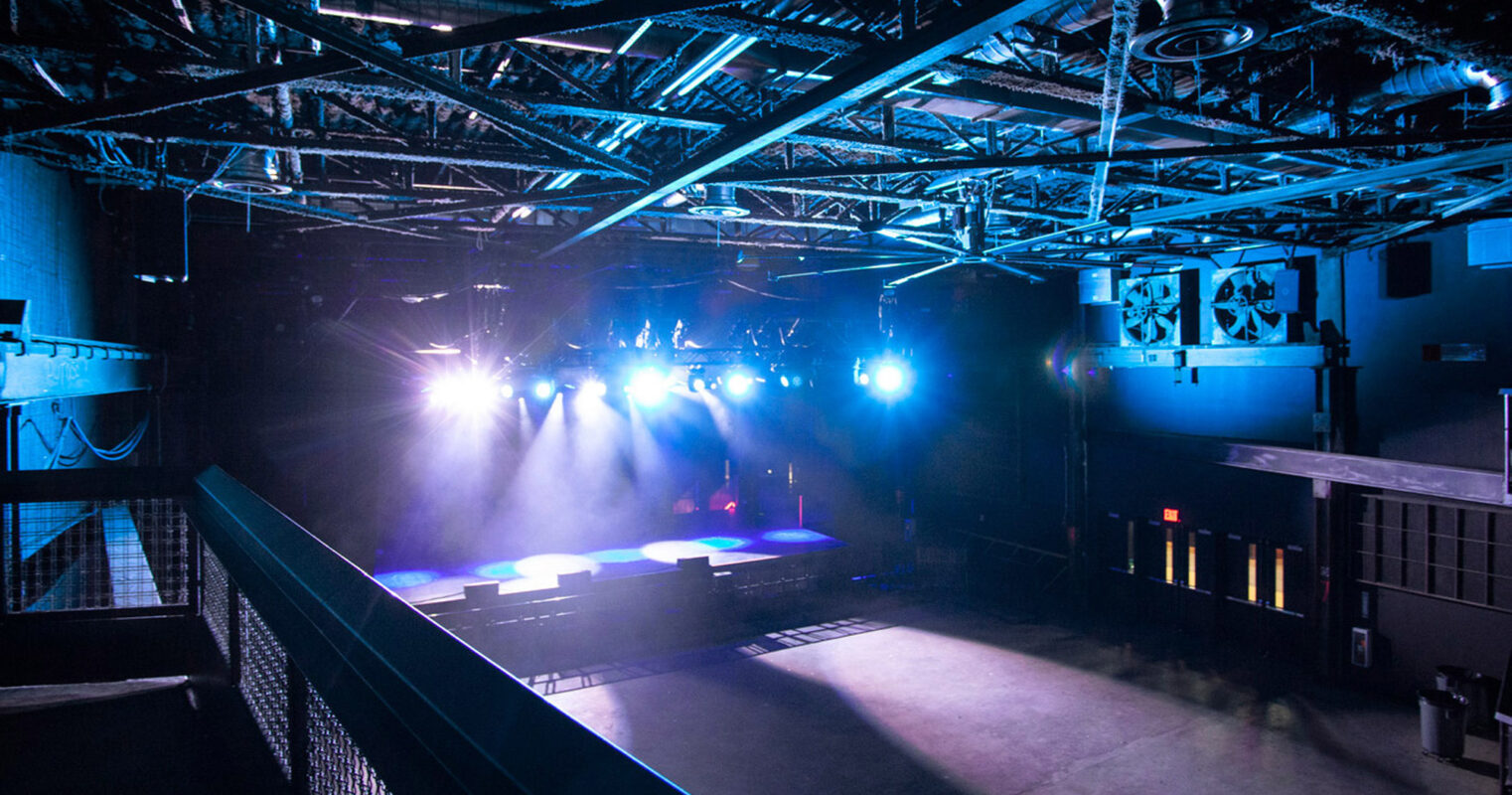 Modern nightclub interior showcasing industrial design elements, with exposed metal trusses, strategic blue and white lighting casting dynamic shadows, and an empty dance floor inviting anticipation of nightly revelries.