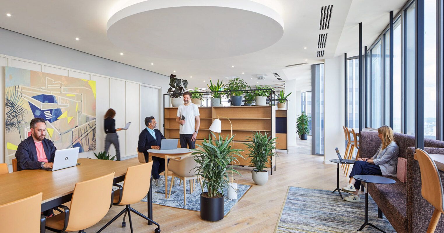 Open-plan office space featuring minimalist wooden desks, ergonomic chairs, and ample natural light from floor-to-ceiling windows. Lush indoor plants provide a refreshing contrast against the sleek, neutral color scheme, reinforcing biophilic design principles.