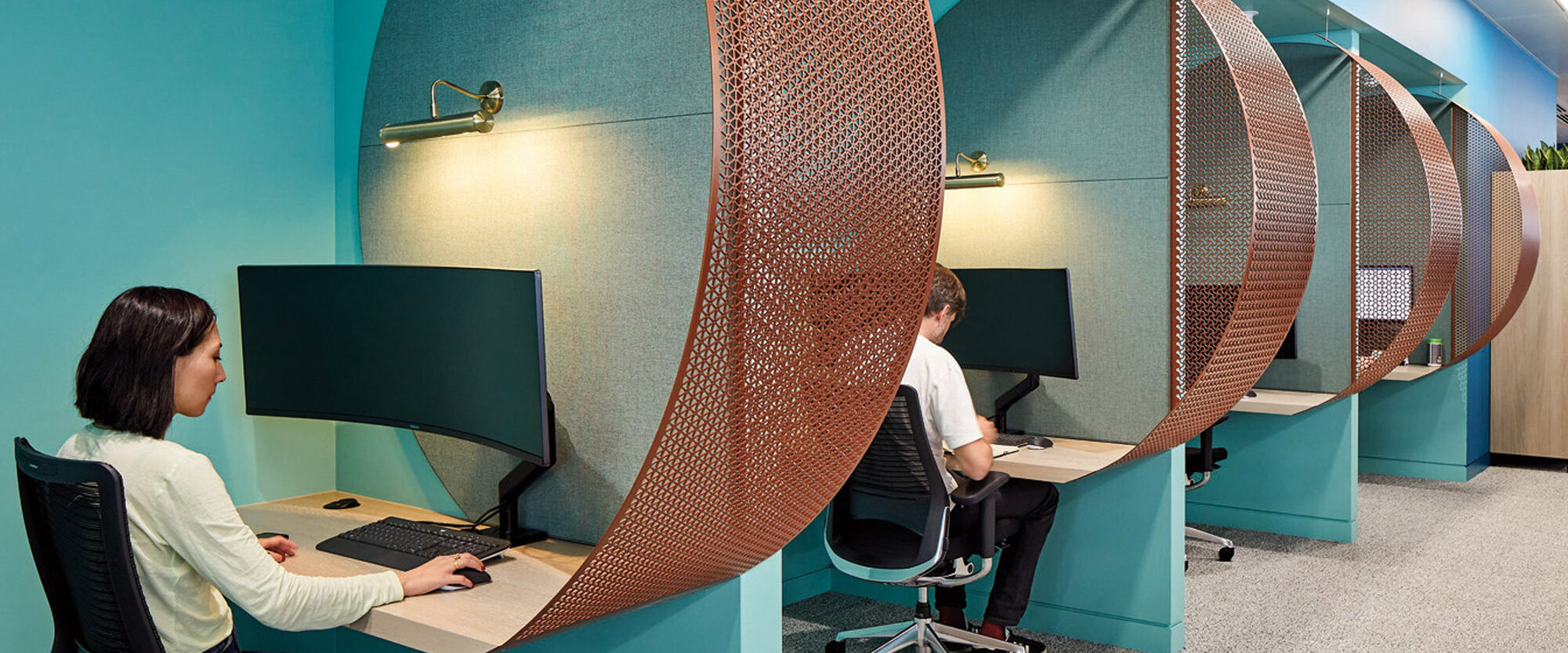 Modern open-plan office featuring turquoise cubicle partitions with curved, copper-finished privacy screens. Ergonomic black office chairs and wooden storage cabinets punctuate the space, showcasing a blend of functionality and contemporary design aesthetics.