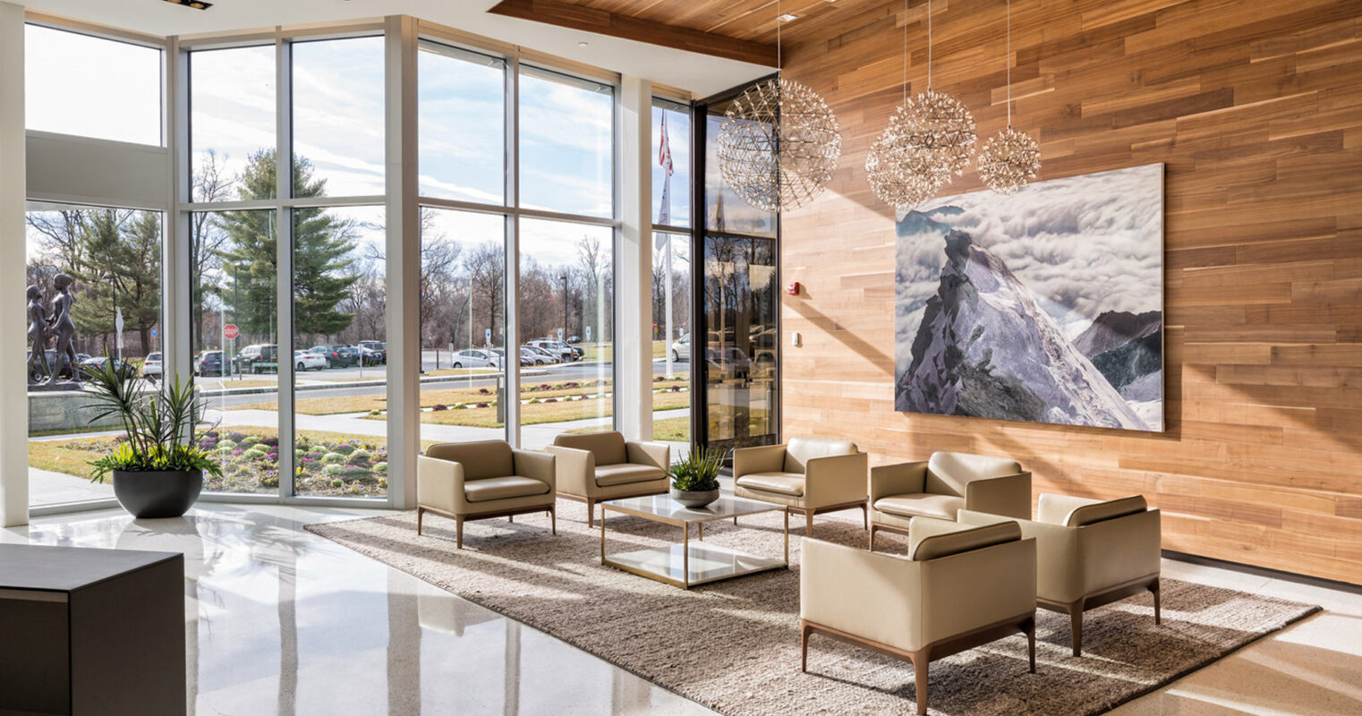 Spacious lobby with floor-to-ceiling windows, modern tan armchairs on a textured rug, wood panel walls, and striking spherical chandeliers. A large mountainous landscape photograph serves as a focal point.