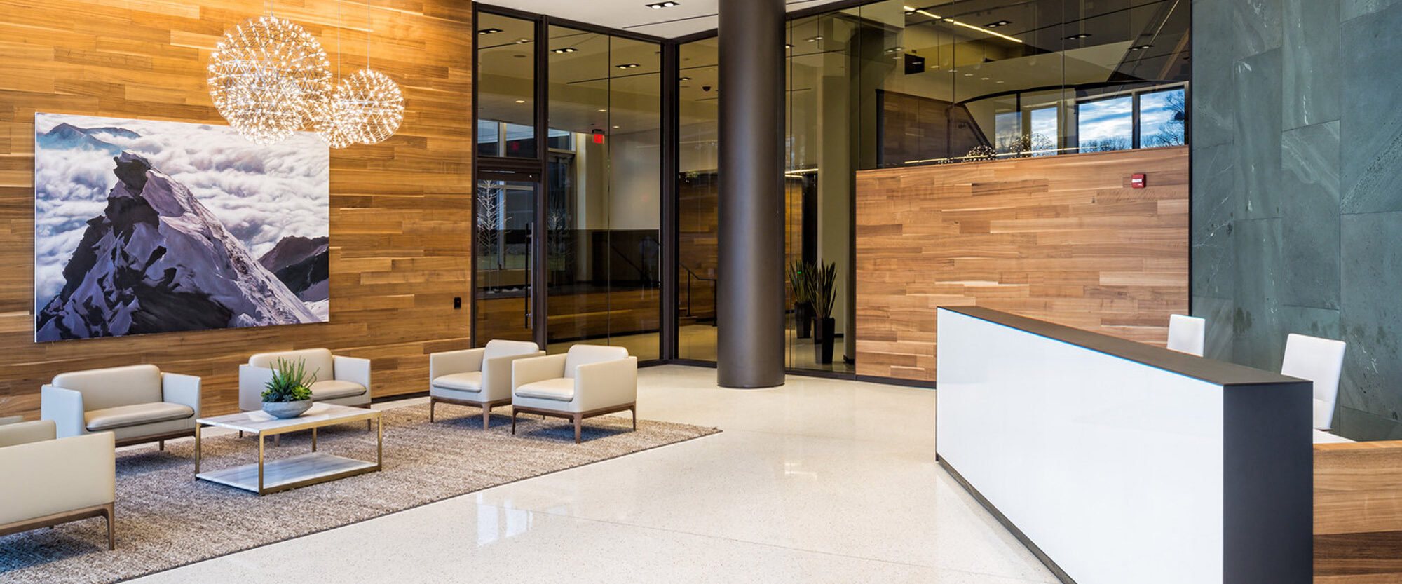 Modern corporate lobby featuring warm wooden wall panels, sleek white reception desk, and contemporary seating arrangement under artistic lighting fixtures, complemented by an oversized mountain landscape painting.