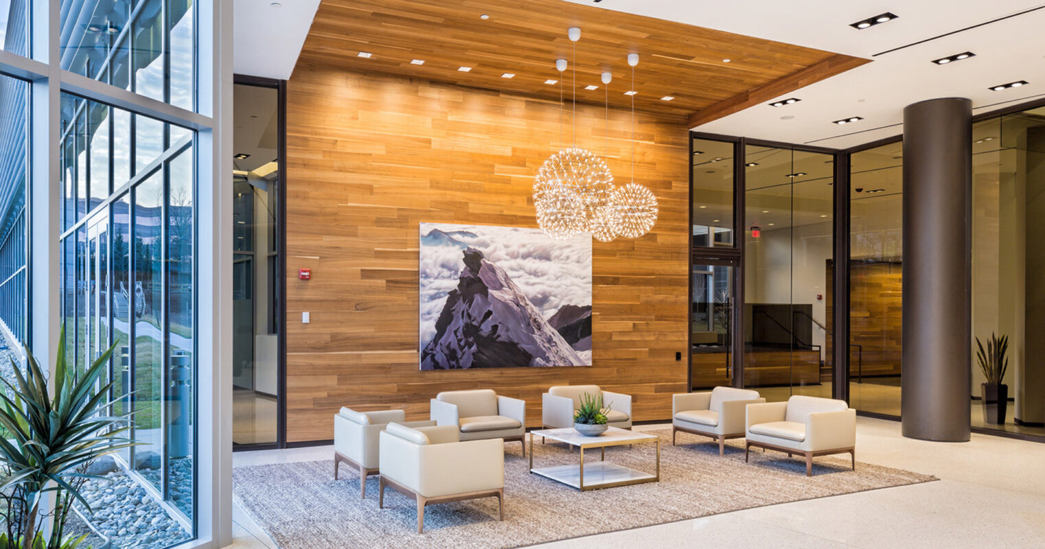 Modern corporate lobby featuring a warm wood accent wall with integrated lighting, flanked by a large mountain landscape artwork. Sleek beige armchairs and minimalist sofas are arranged on a neutral-toned rug, beneath spherical pendant lights, creating a welcoming atmosphere.