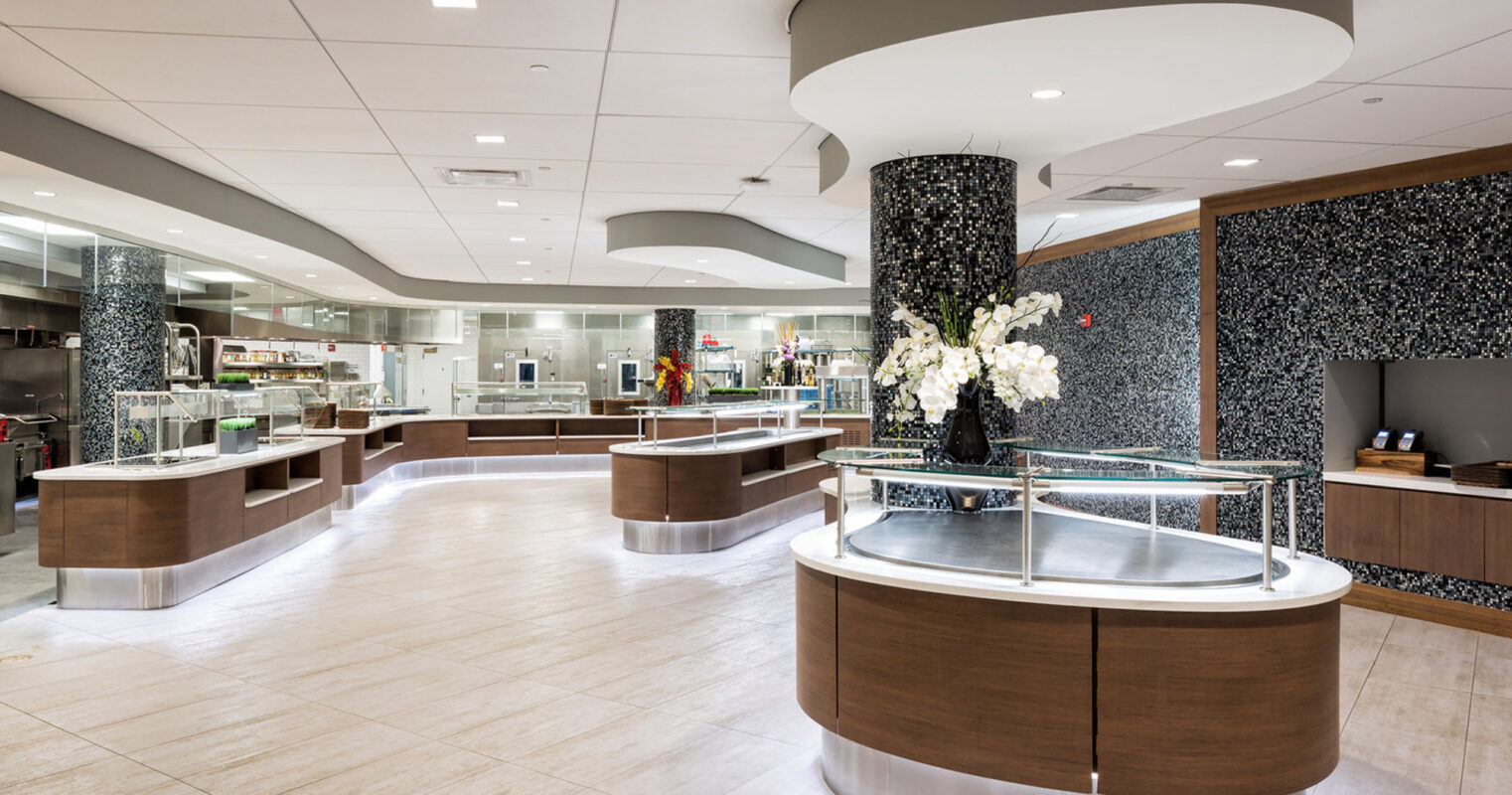 Modern commercial kitchen with curved walnut reception desks, black granite countertops, and cylindrical accents. White orchids add elegance against a black stone-tiled column, complemented by recessed and pendant lighting in the open, neutral-toned space.