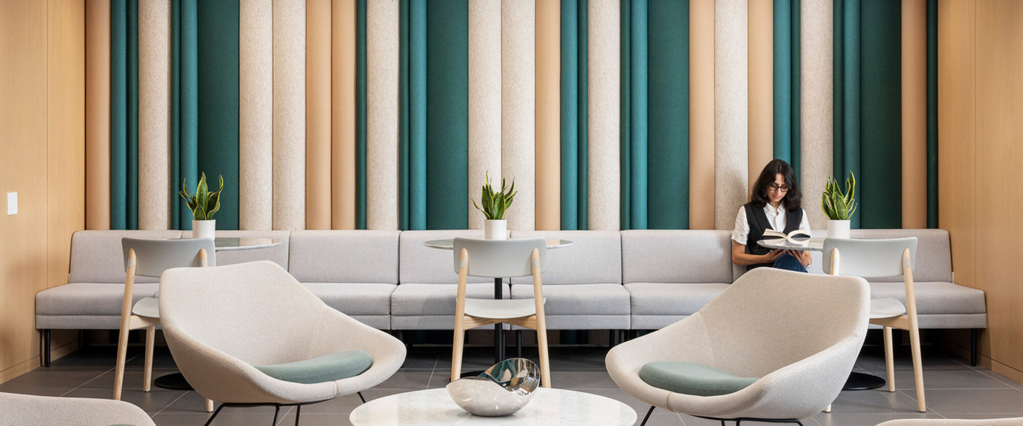 Contemporary waiting area with varied textures; soft beige couches complement wall panels in muted greens and creams. Overhead, a sleek white slatted ceiling contrasts the warm wooden floor. Central tables and modern lounge chairs offer a relaxed space for reading and work.