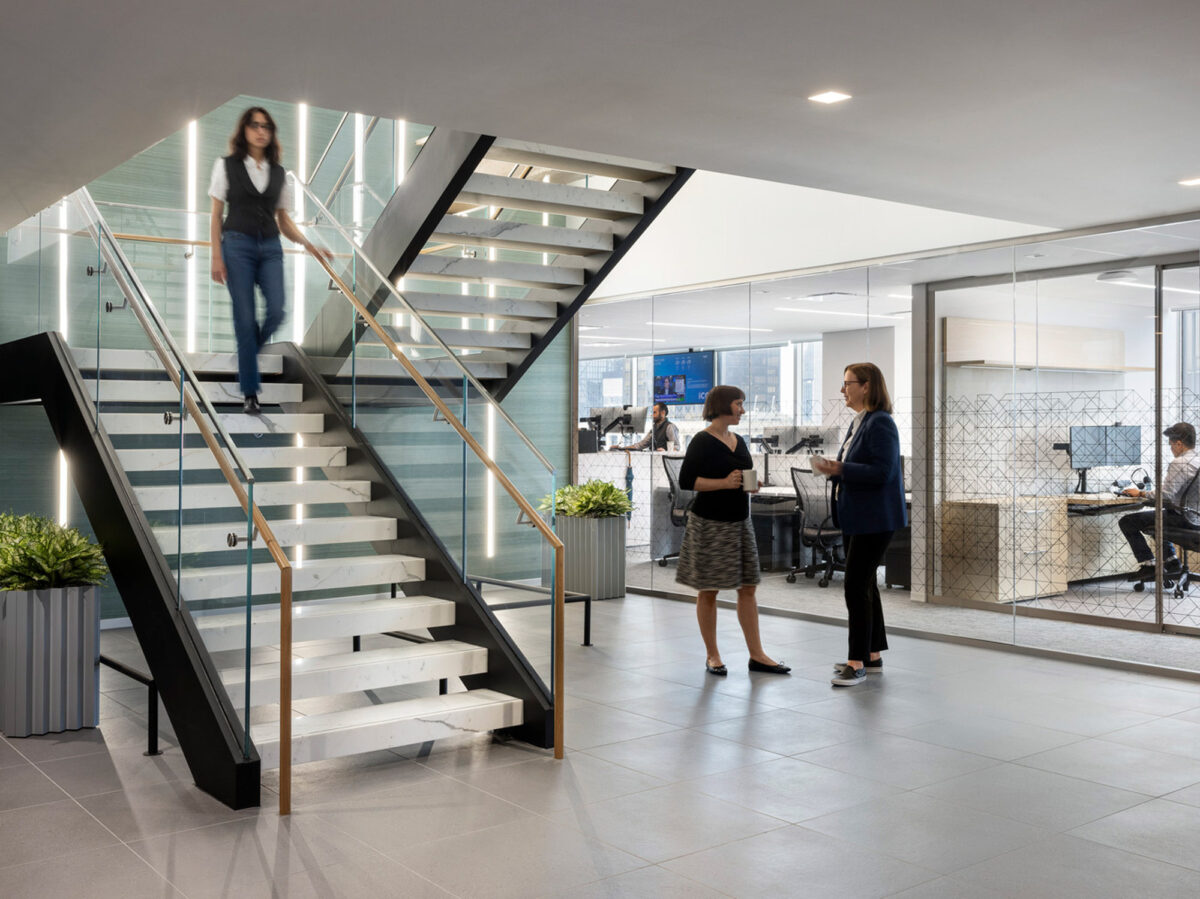 Modern office space featuring a sleek glass and steel staircase with floating treads, connecting two levels. Employees engage in conversation near the base, with open-concept workspaces visible in the background. The design fosters transparency and encourages collaboration.