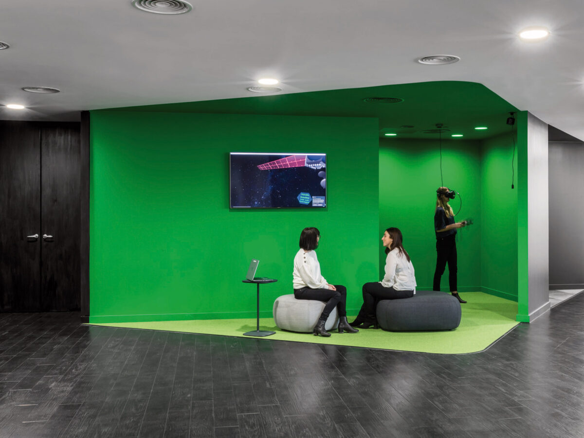 This office lounge features a striking green accent wall with mounted television, contrasted by subdued grey flooring and ceiling. Employees engage in a relaxed setting, enhancing the modern workspace environment with a blend of bold color and minimalist furniture design.