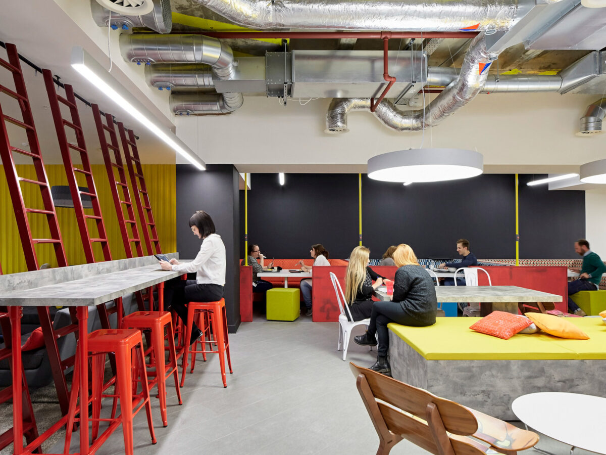 Modern office break room featuring exposed industrial ceiling, bright red structural elements, white pendant lighting, and a mix of bar-height and lounge seating with vibrant pops of yellow and green.