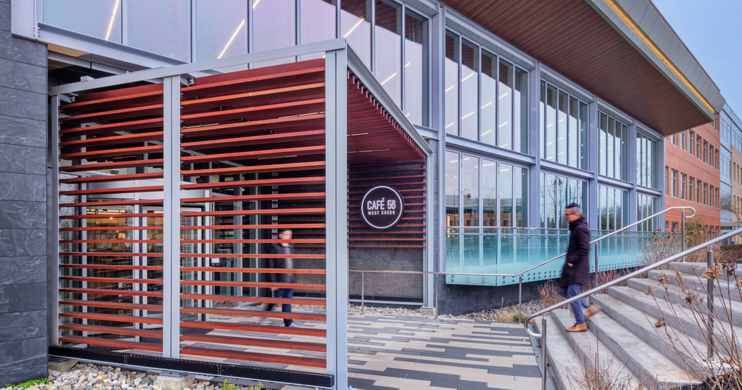 Modern cafe exterior featuring a combination of stone, wood, and metal materials with slatted wooden screens, large glass windows, and a prominent overhanging roof with geometric details, set against a landscaped entrance with concrete steps.