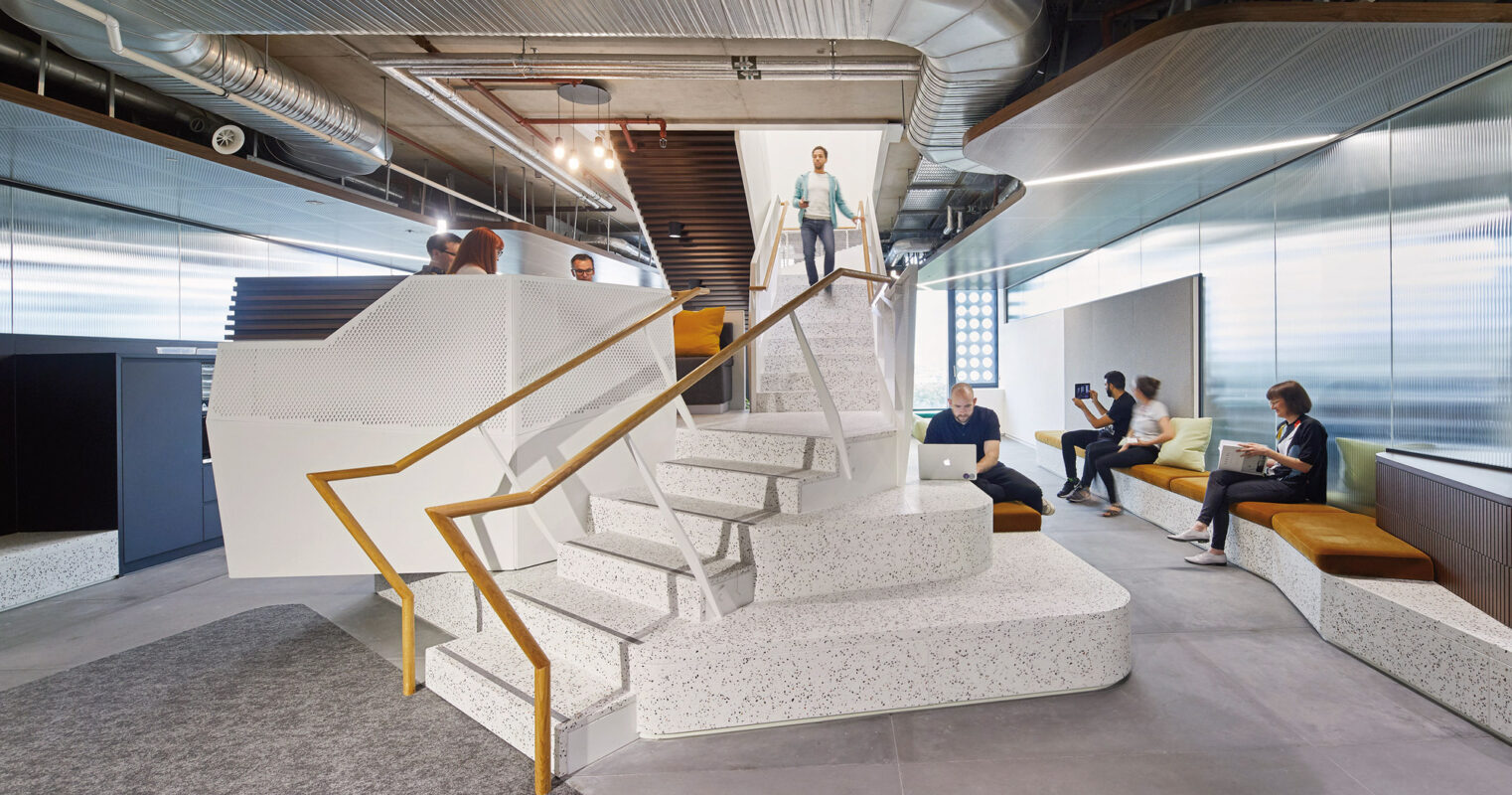 Modern office interior featuring a central floating staircase with white steps and gold accents. The surrounding open-concept space includes a lounge area with mustard yellow seating, exposed ductwork overhead, and people engaged in casual conversation, embodying a contemporary work environment.