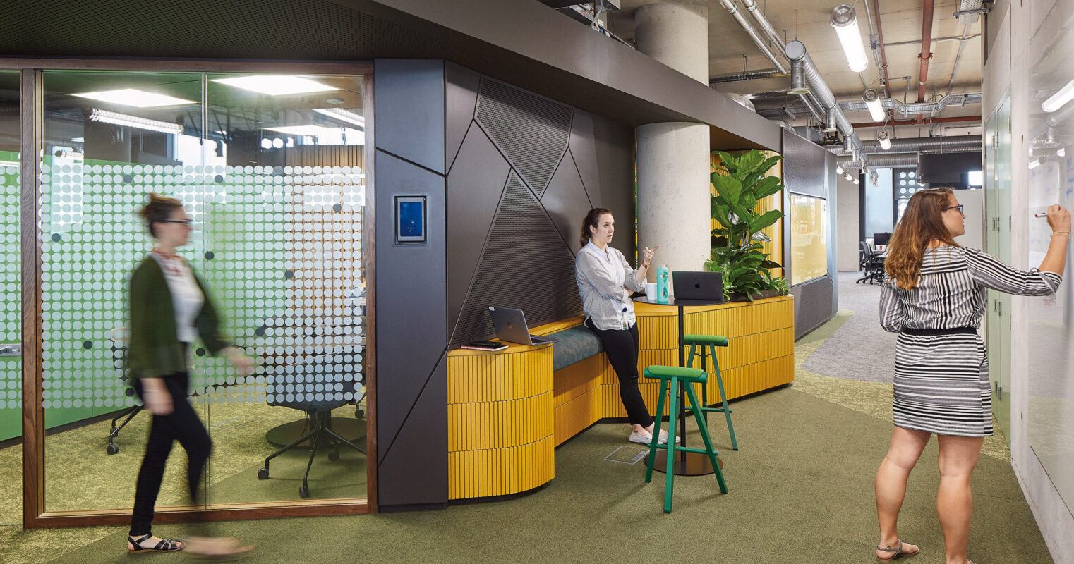 Modern office lobby showcasing geometric gray wall paneling, a reception desk with dynamic yellow accents, and high green stools. Dot-patterned glass partitions enhance privacy, while an employee interacts with a wall-mounted interface opposite to a glass whiteboard in use.