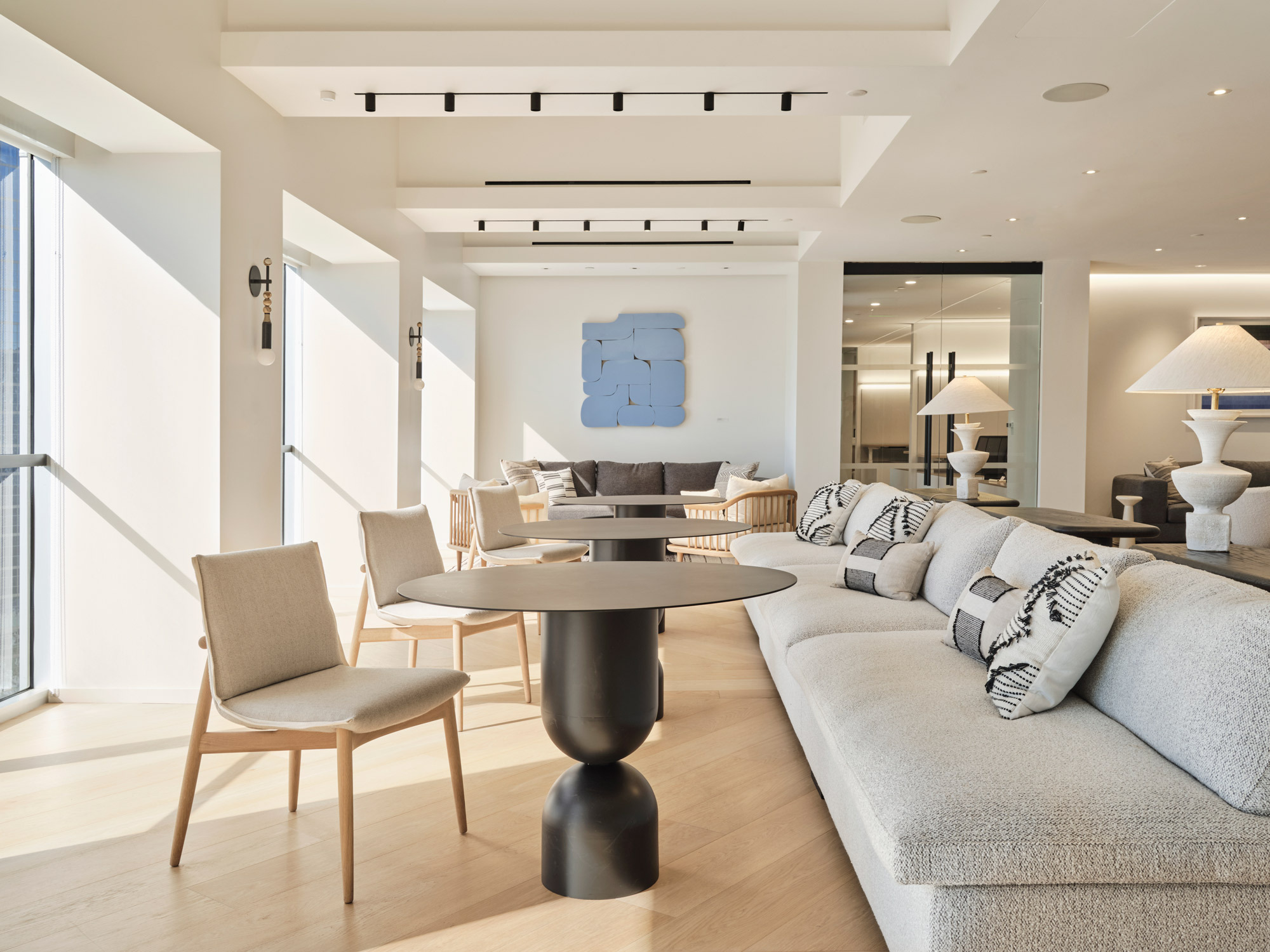 Spacious living room with abundant natural light, featuring a minimalist color palette. Sleek, low-profile furniture with neutral upholstery complements the light wooden flooring and white beam ceiling, accented by a bold blue abstract wall art.