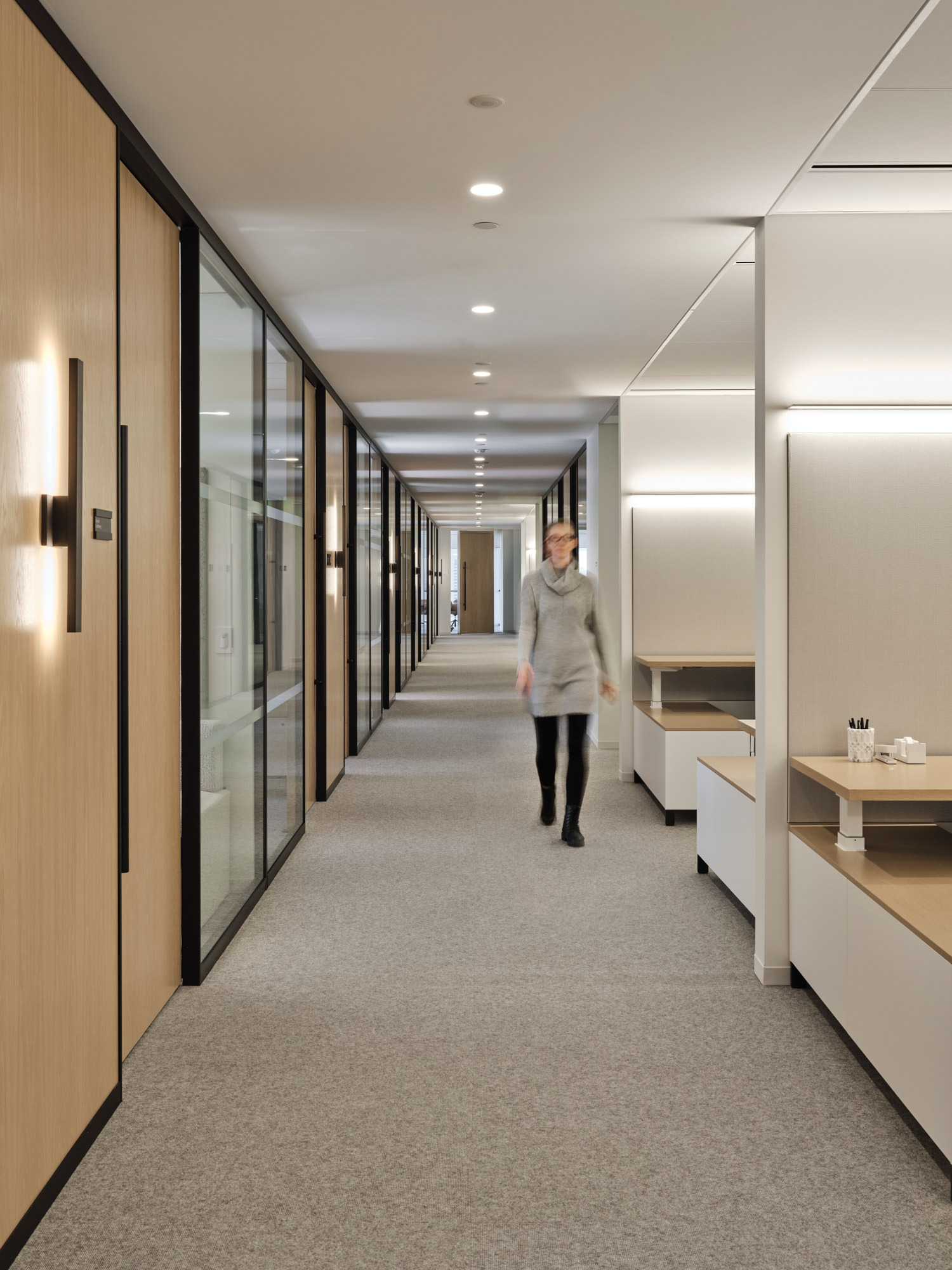 Modern office corridor featuring natural wood-framed glass partitions, recessed linear lighting, and textured grey carpeting. A person strides through, illustrating the space's human scale and flow. The design balances warmth and professionalism.