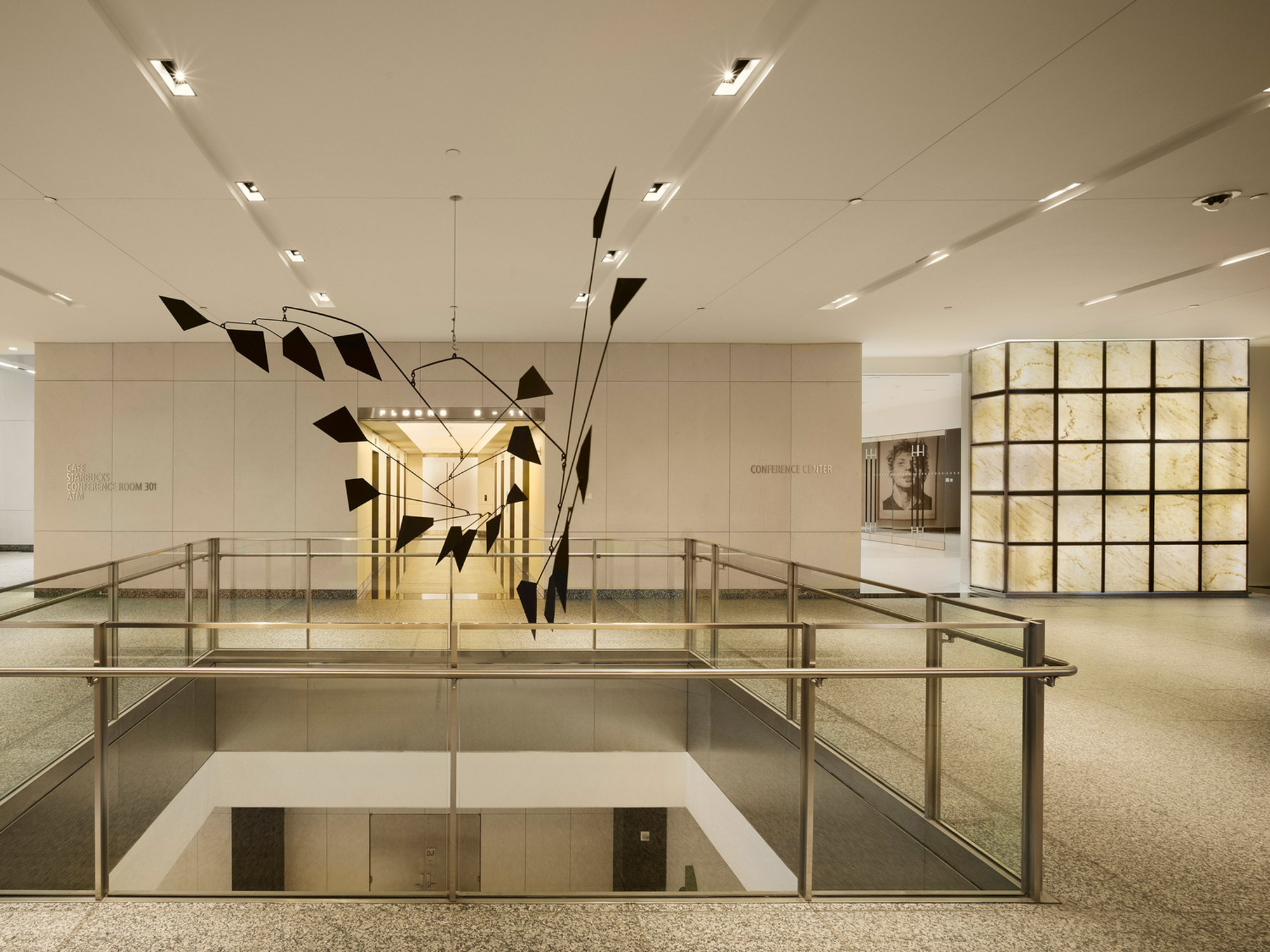 Minimalist lobby featuring a mobile sculpture with geometric shapes suspended above, complemented by warm, diffused lighting from a backlit translucent wall panel and the use of reflective materials for an elegant, modern atmosphere.