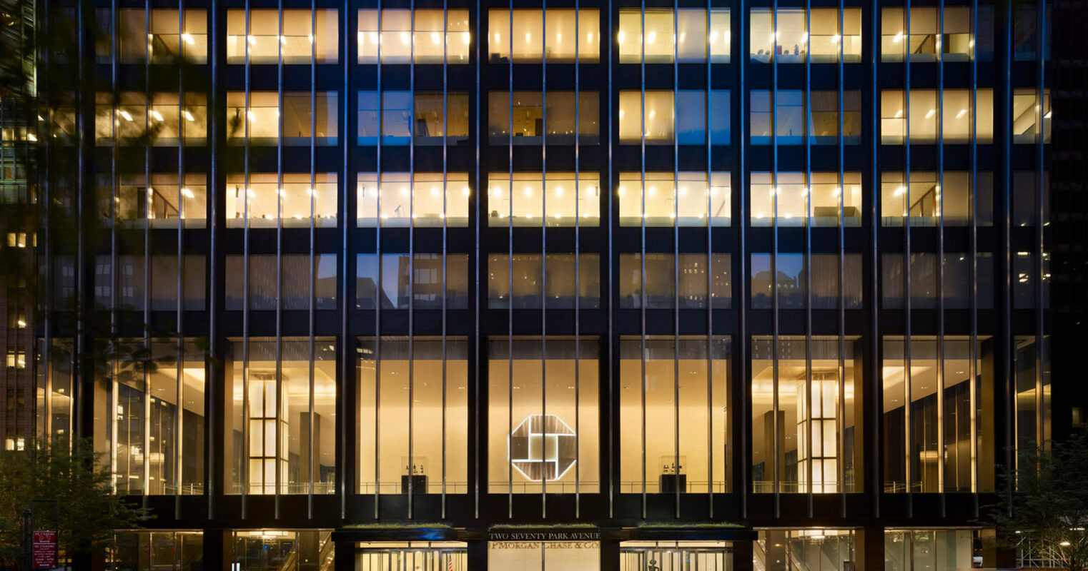 Modern office building facade illuminated at twilight, showcasing symmetrical windows with warm interior lighting, and a grand entrance flanked by sleek columns.