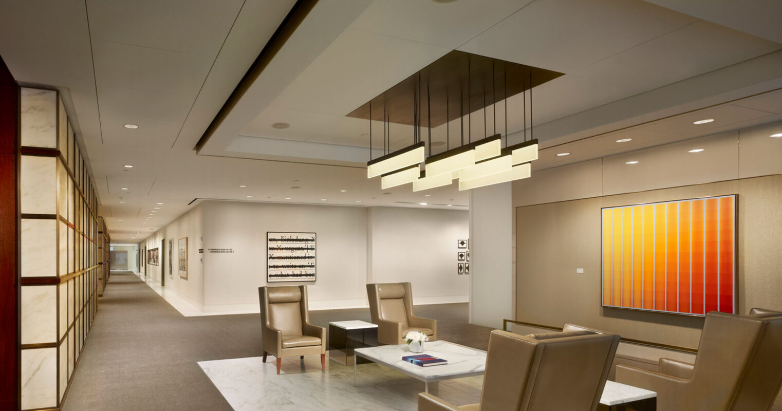 Minimalist office lobby with sleek, neutral-toned furniture, white marble floors, and linear lighting fixtures. Contemporary art adorns the walls, and a drop-down wooden accent panel adds warmth to the space.