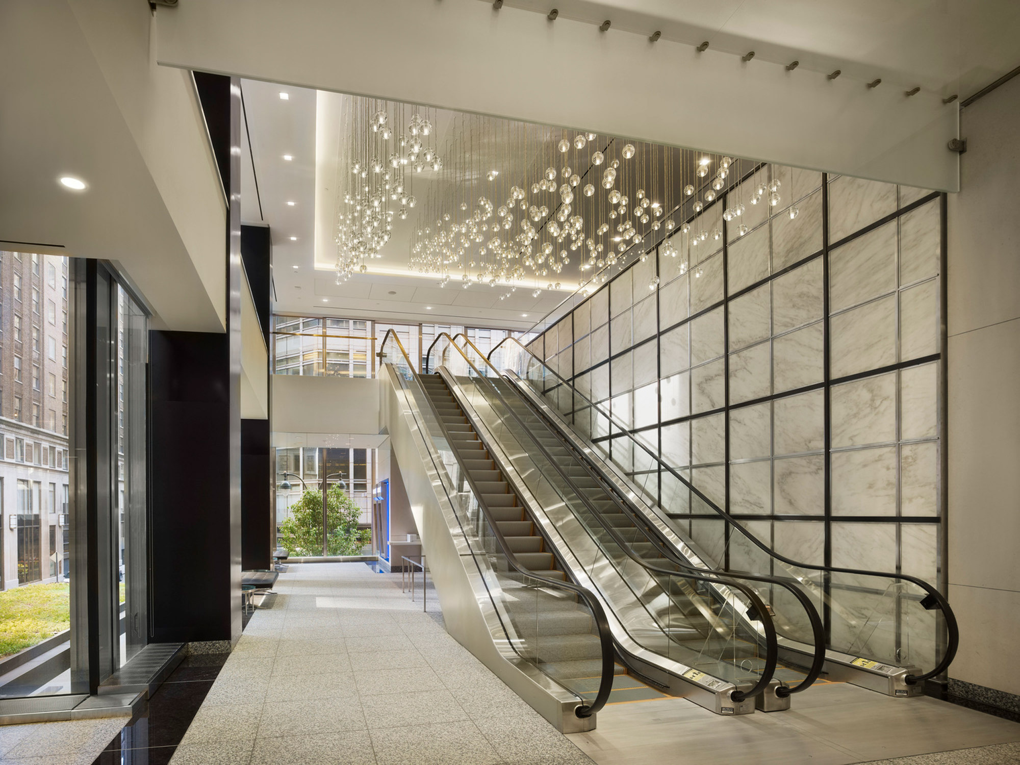 Luminous modern lobby with a pair of sleek escalators, illuminated by a floating bubble-like chandelier. The backlit translucent stone wall panels exude a warm glow, enhancing the luxurious ambience.