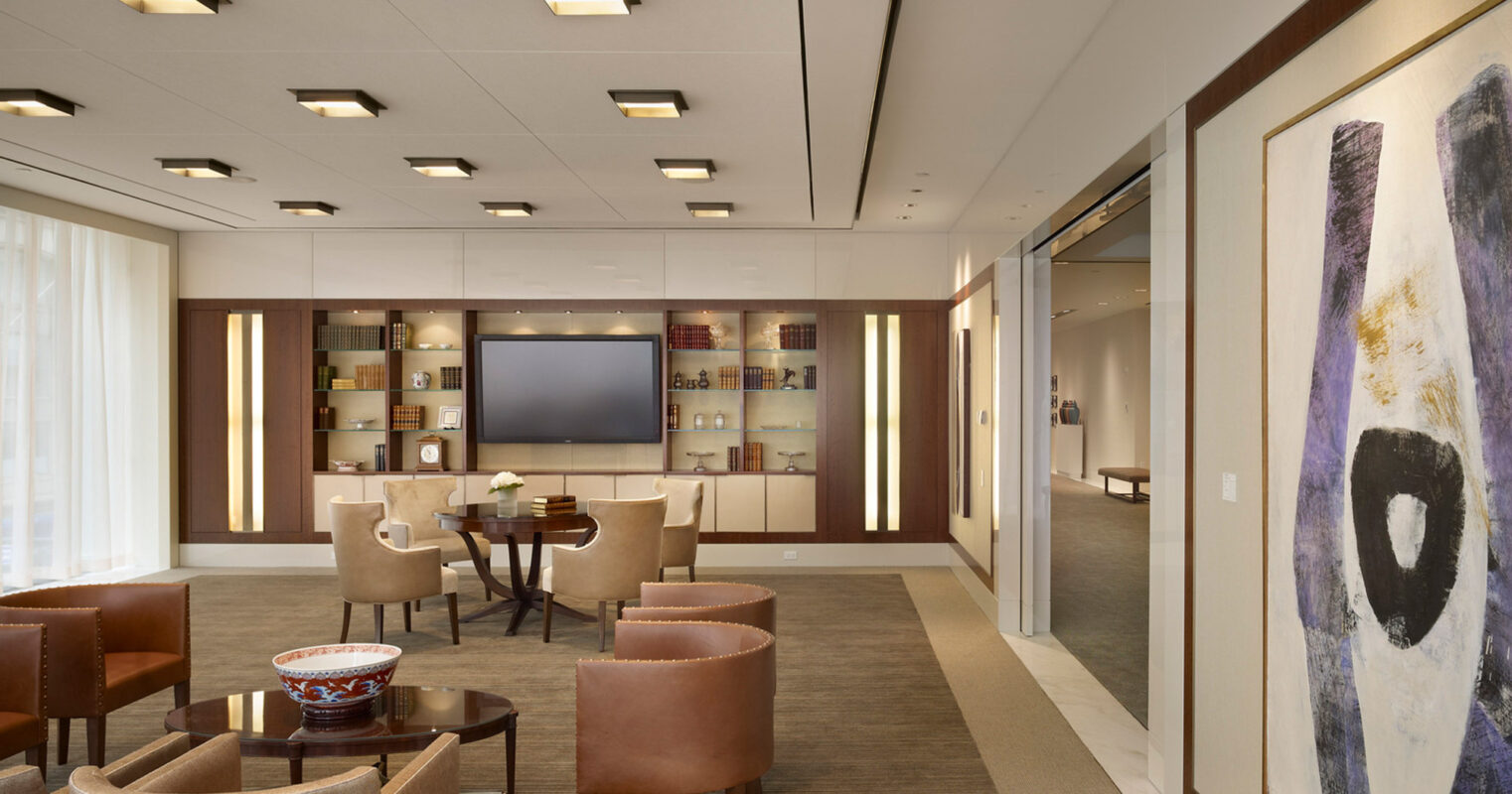 Contemporary office lounge with neutral tones, featuring armchairs around a central coffee table, recessed lighting in a grid pattern, and an abstract wall painting complementing built-in shelving and glass partitions.