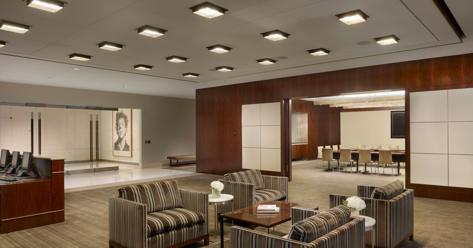 Modern corporate lobby featuring striped upholstered seating, recessed ceiling lights, dark wood paneling, and neutral-toned carpet. Glass partitions provide a glimpse into the adjacent conference room, creating a sense of openness while preserving privacy.