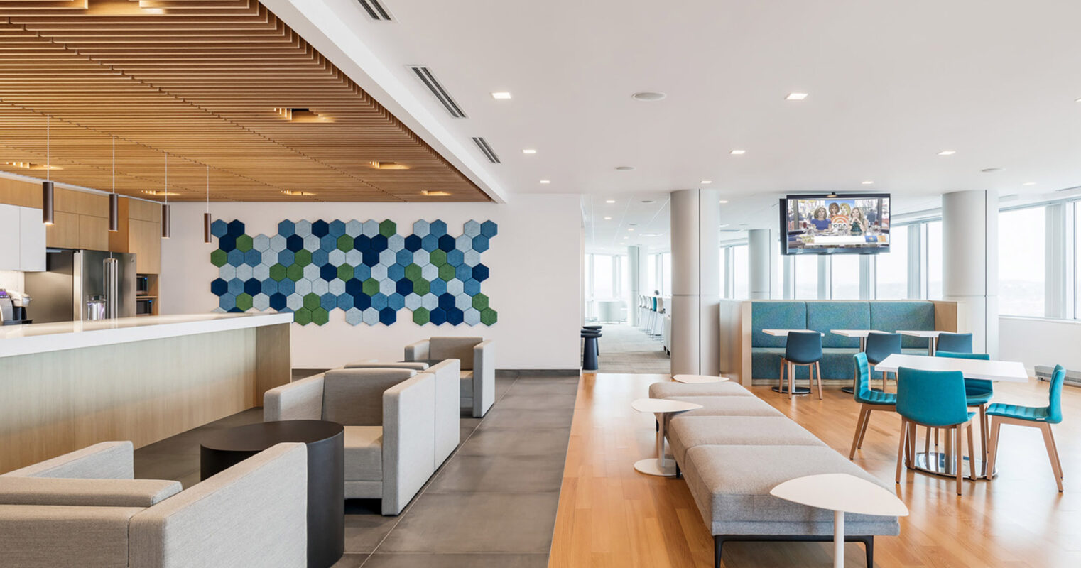 Modern office breakroom featuring natural light, sleek white surfaces, wooden slats ceiling detail, and pops of color from blue upholstered furniture and hexagonal wall art.