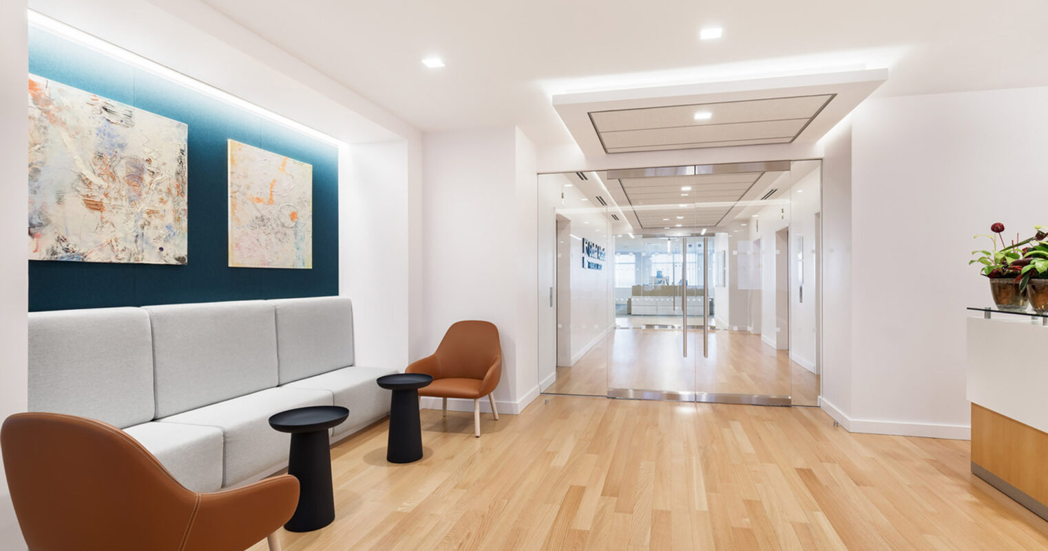Spacious hallway featuring light hardwood flooring, white walls, and recessed ceiling lighting. A minimalist seating area with a gray sofa, vibrant abstract art, and brown accent chairs contrasts with the clinical glass-door entryway at the end of the corridor.